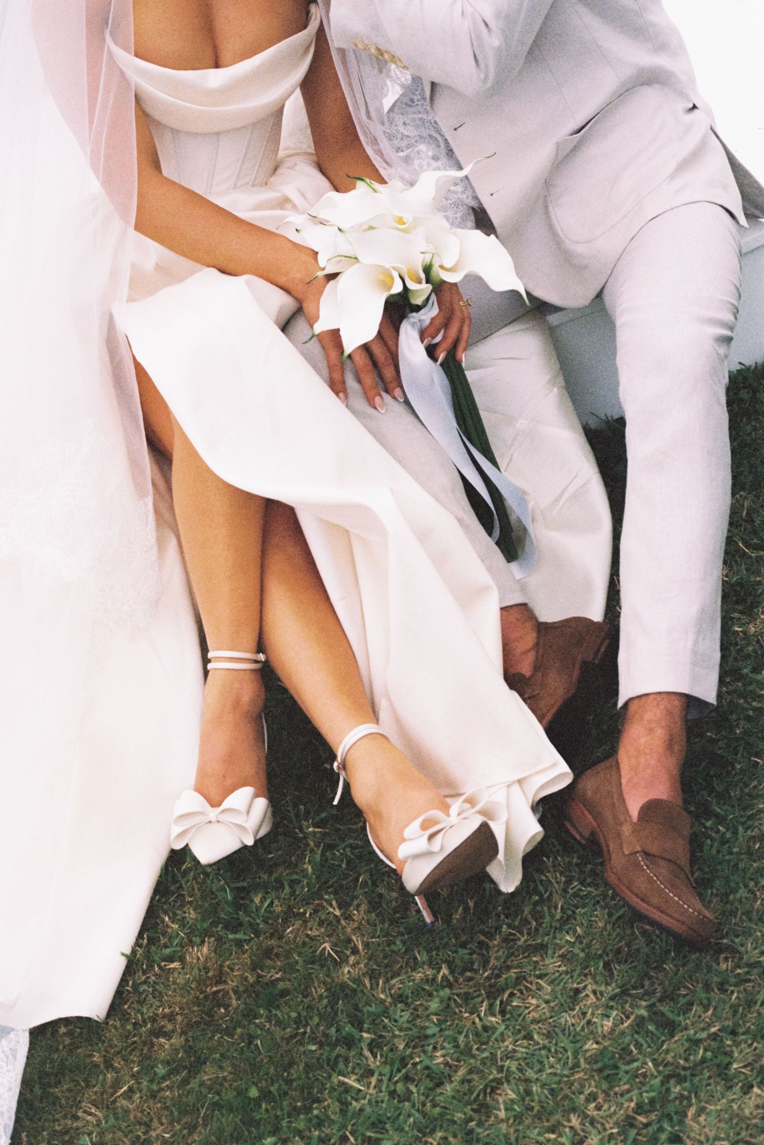 A row of people sitting on grass wearing white formal attire and dress shoes at an outdoor event.