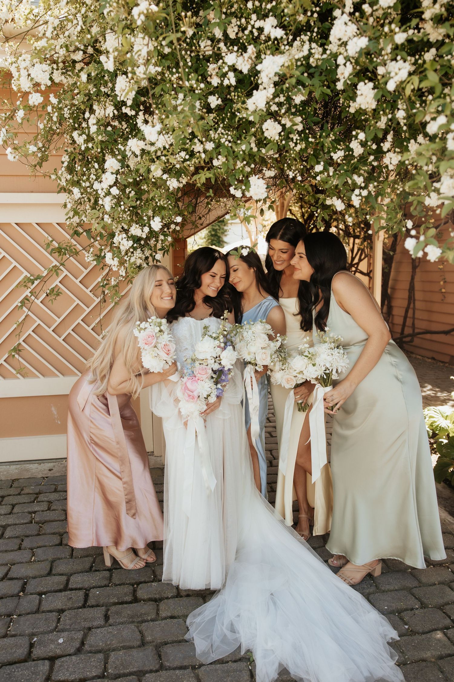 A bridal party in pastel dresses laughs together under a white floral archway in an outdoor courtyard setting.