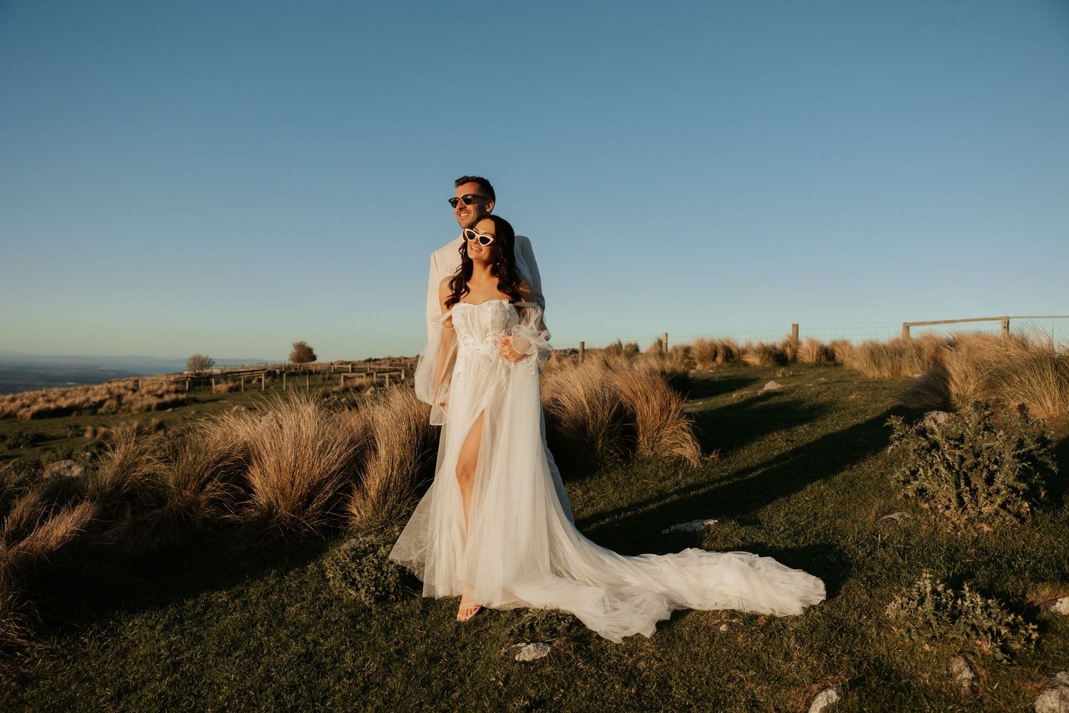 A couple embraces on hilltop at sunset with flowing white wedding dress against golden grass and blue sky.