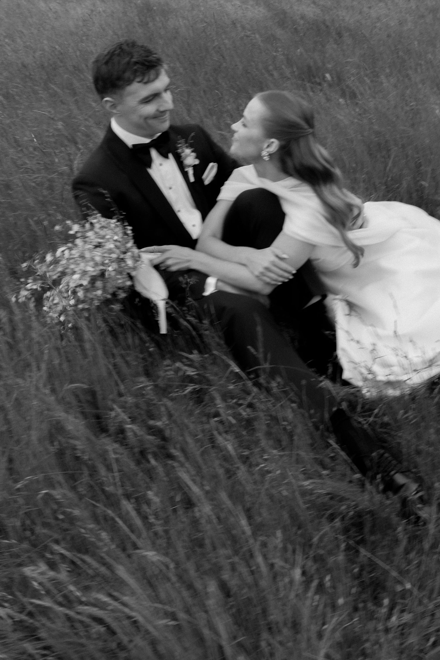 Black and white photo of a bride and groom sitting in tall grass, smiling at each other on their wedding day.