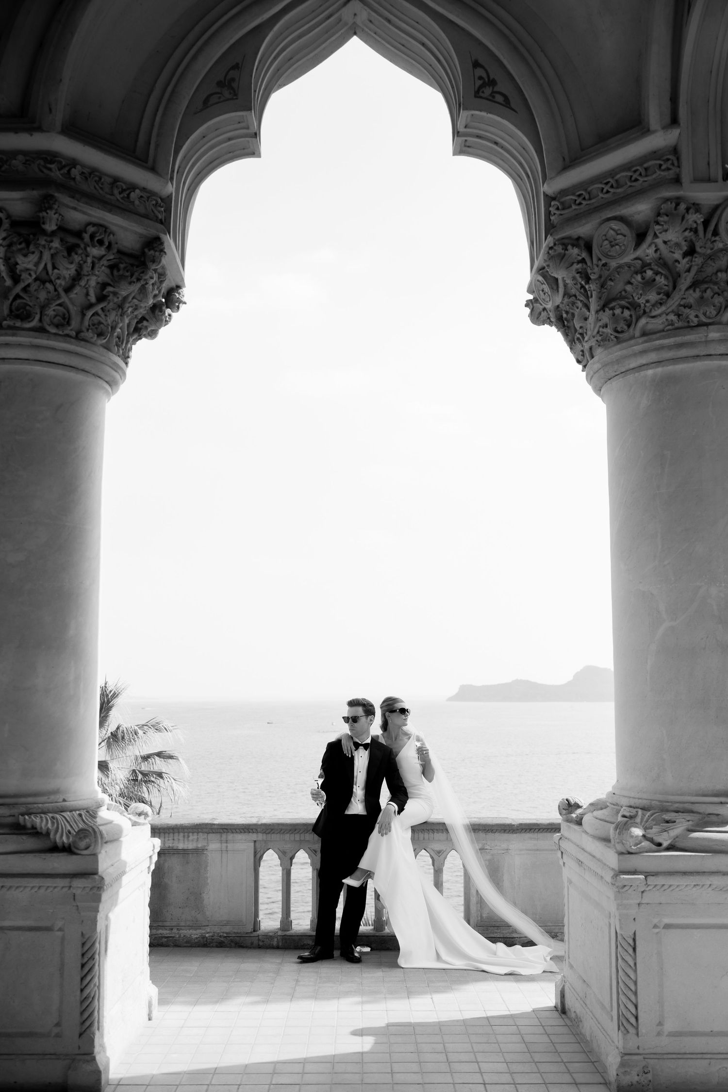 Black and white wedding photo of bride and groom posing under ornate arch with ocean view backdrop.