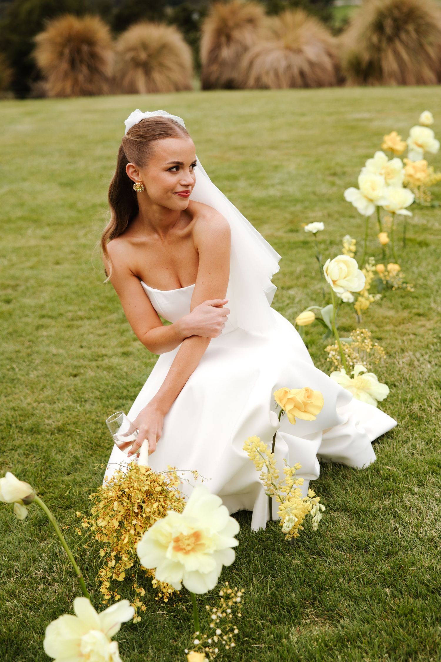 Bride in white strapless gown sitting on green grass surrounded by yellow and white flowers outdoors.