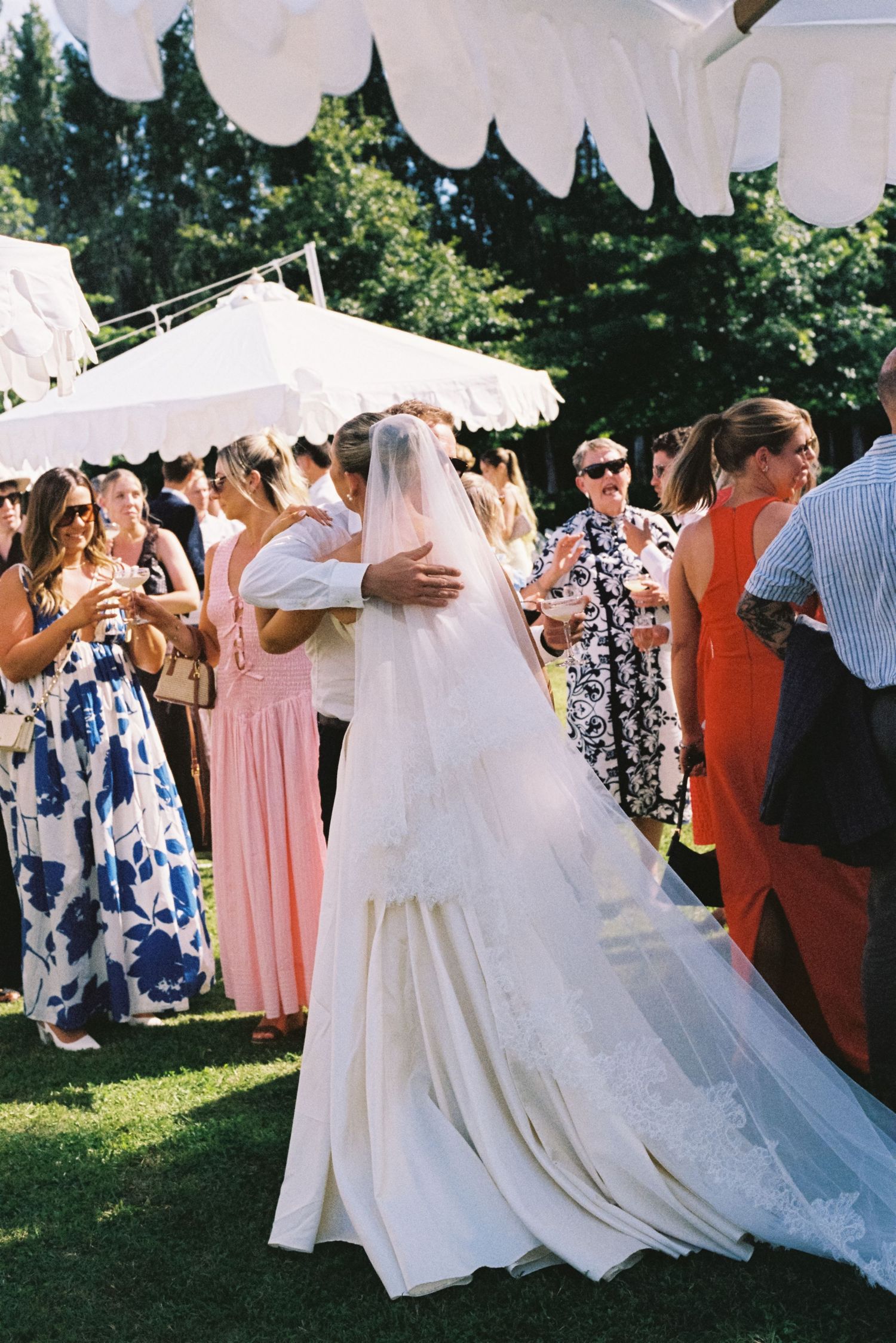 A group of wedding guests in blue and pink dresses dance in a line under white tent decorations outdoors.