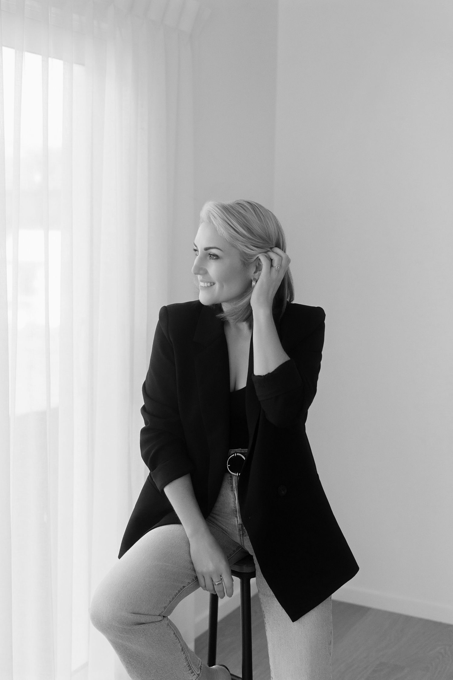 Black and white portrait of a smiling woman in a black blazer and light jeans sitting on a stool beside sheer curtains.