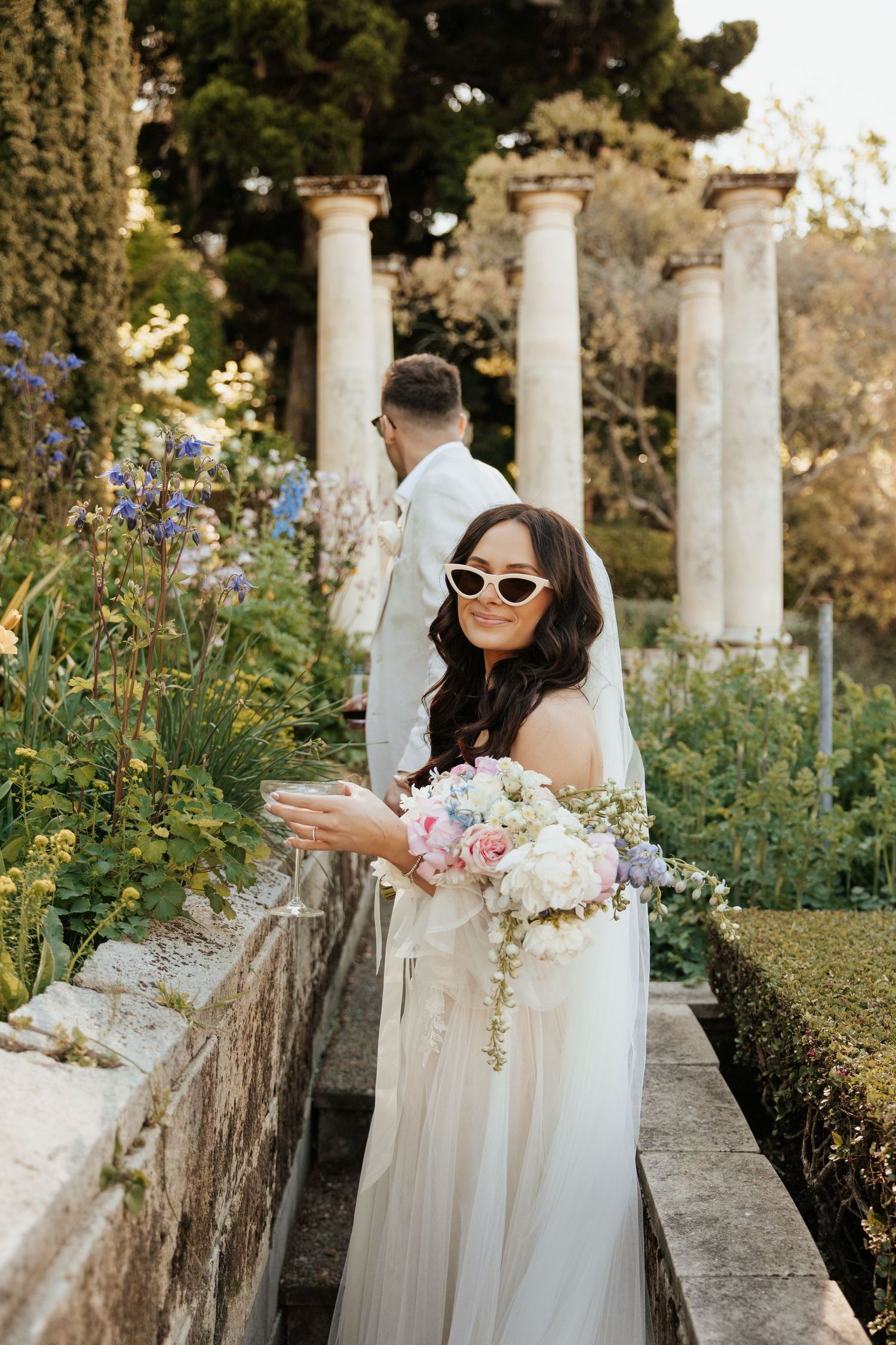White-dressed figure holding a pink bouquet poses in sunglasses by classical stone columns in a garden setting.
