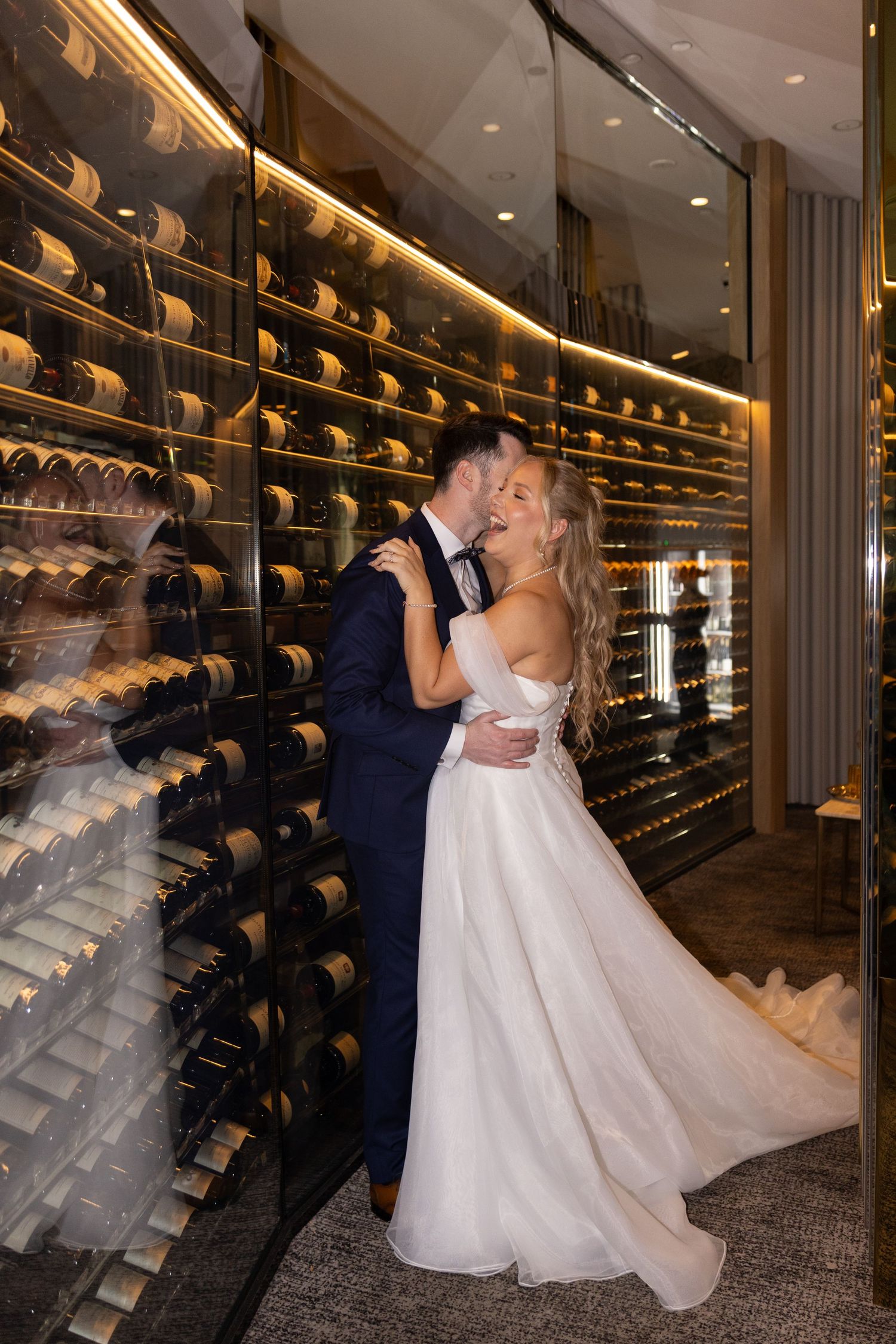 Bride and groom share a romantic kiss in an elegant wine cellar, surrounded by floor-to-ceiling bottle displays.