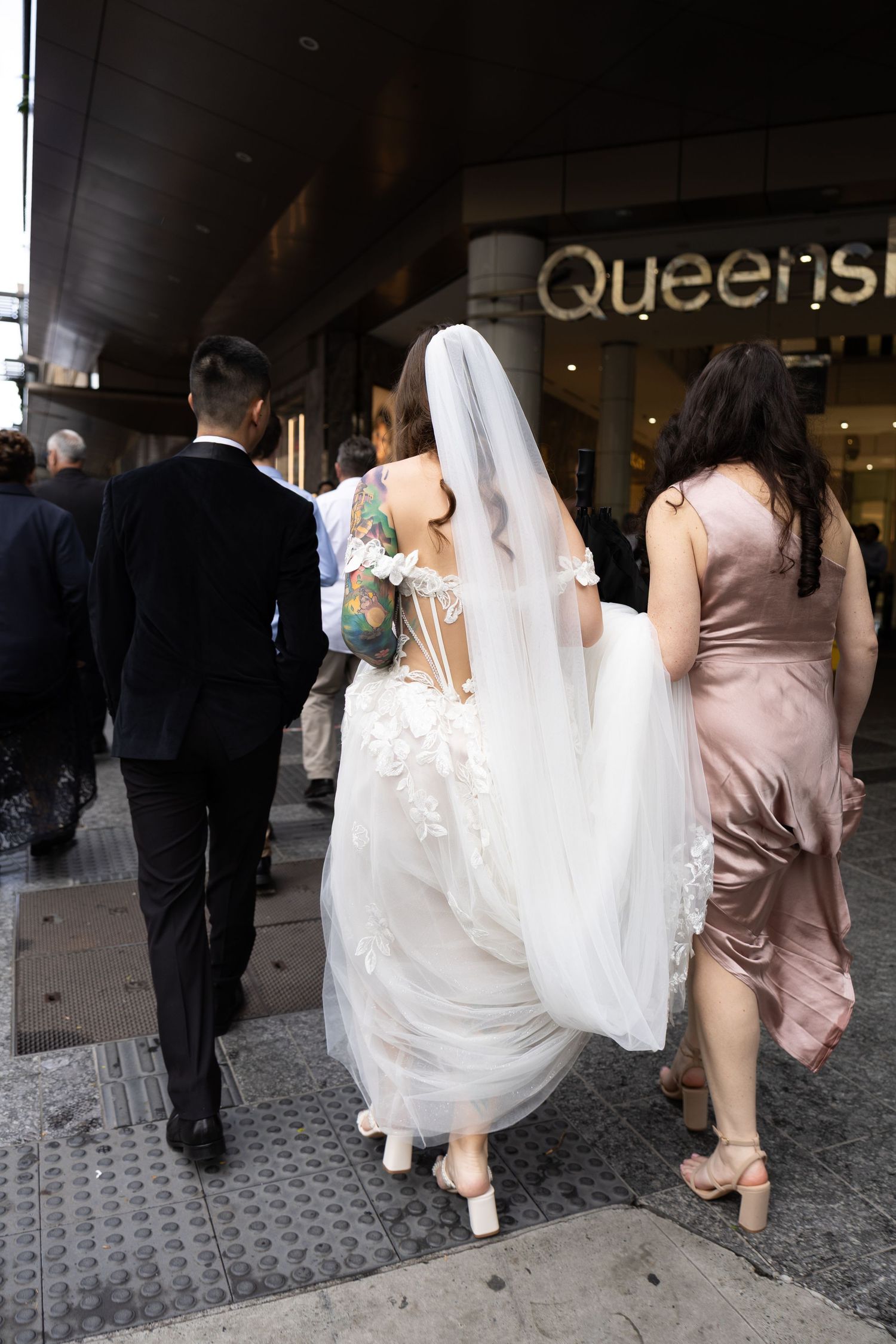 Bride in white gown and veil walks with wedding party on city sidewalk outside Queens venue.