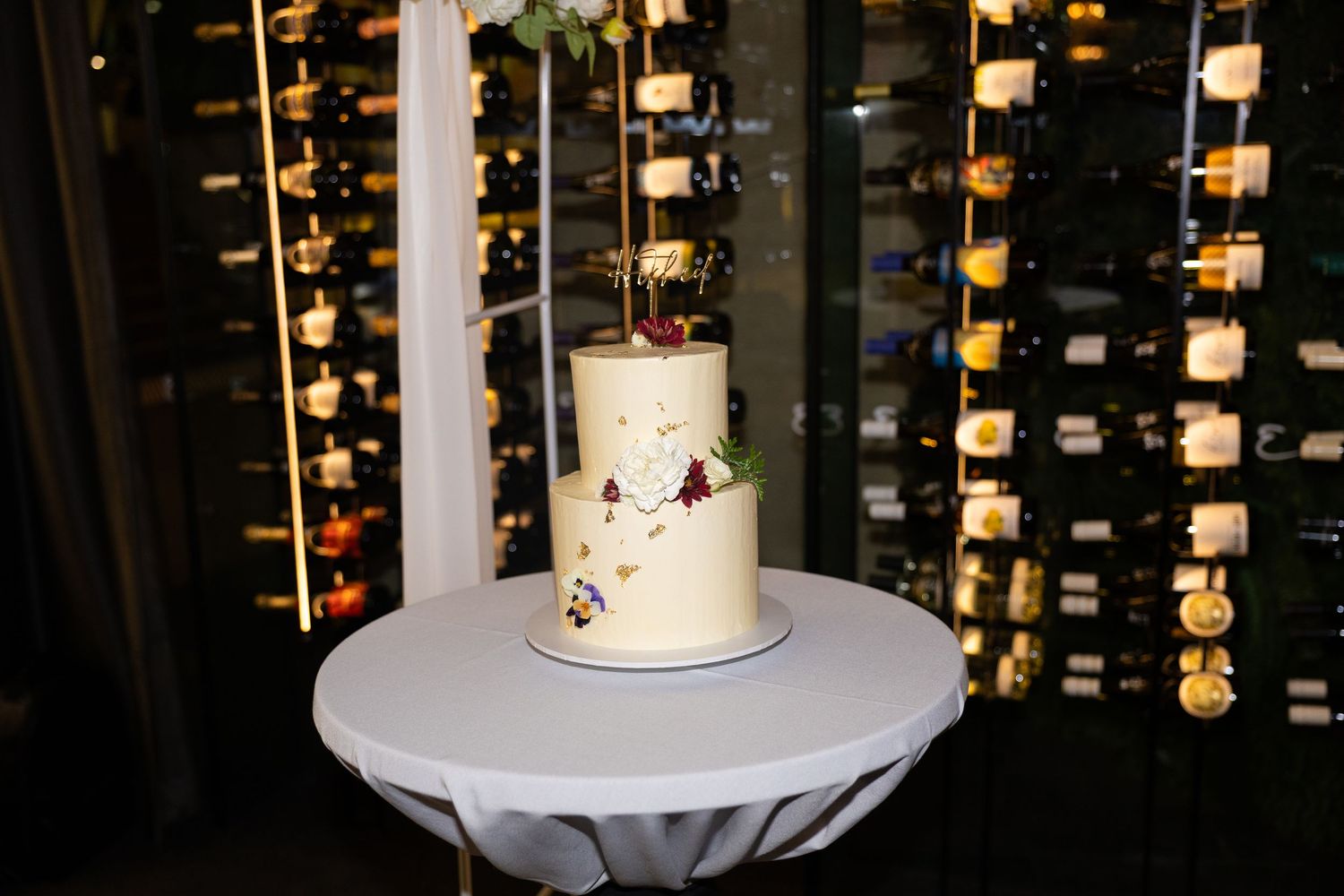 Elegant two-tier white wedding cake with floral accents displayed on a round table in front of a wine cellar wall.