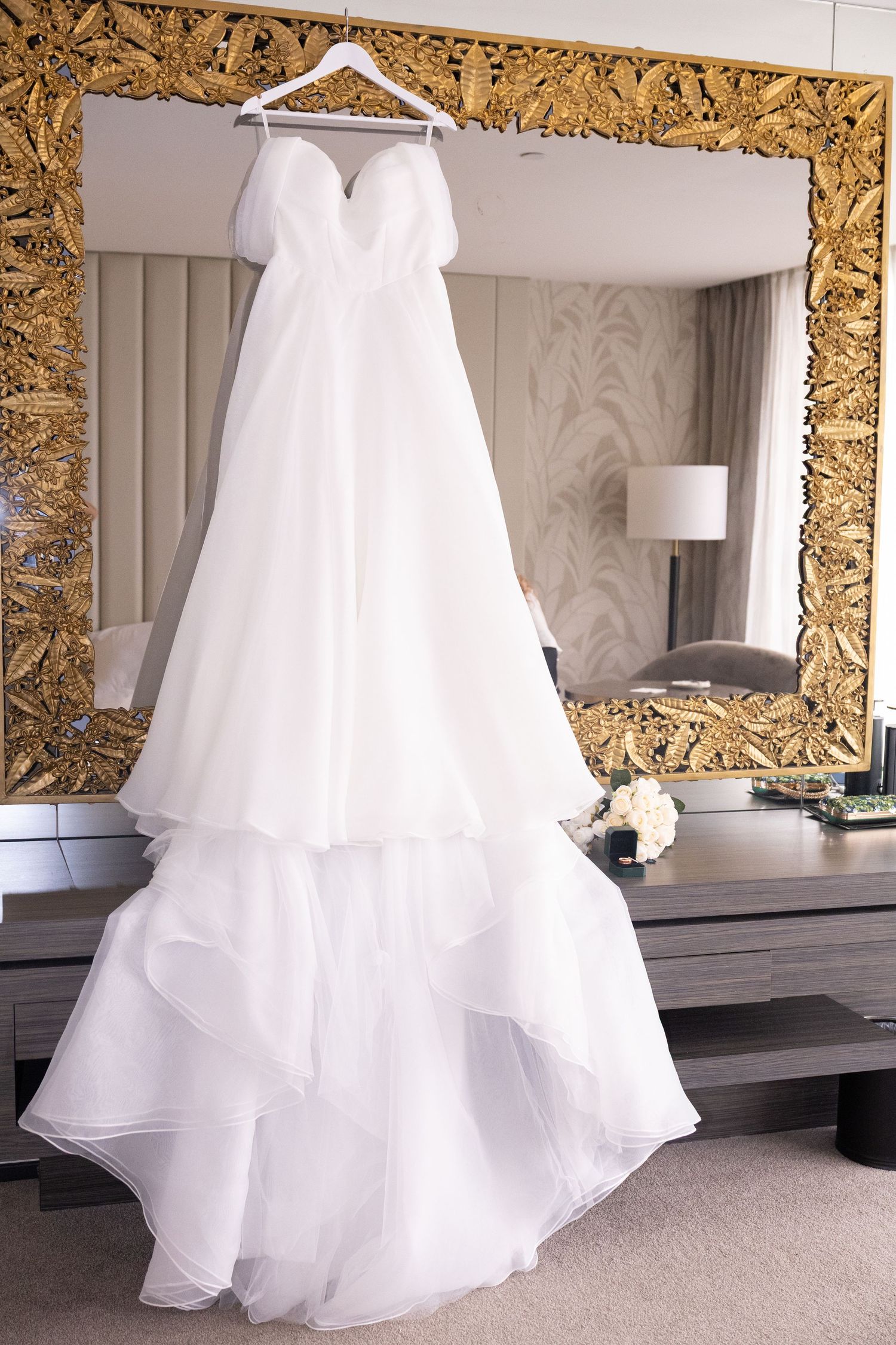 White strapless layered wedding gown hanging on ornate gold mirror frame in elegant hotel bridal suite.
