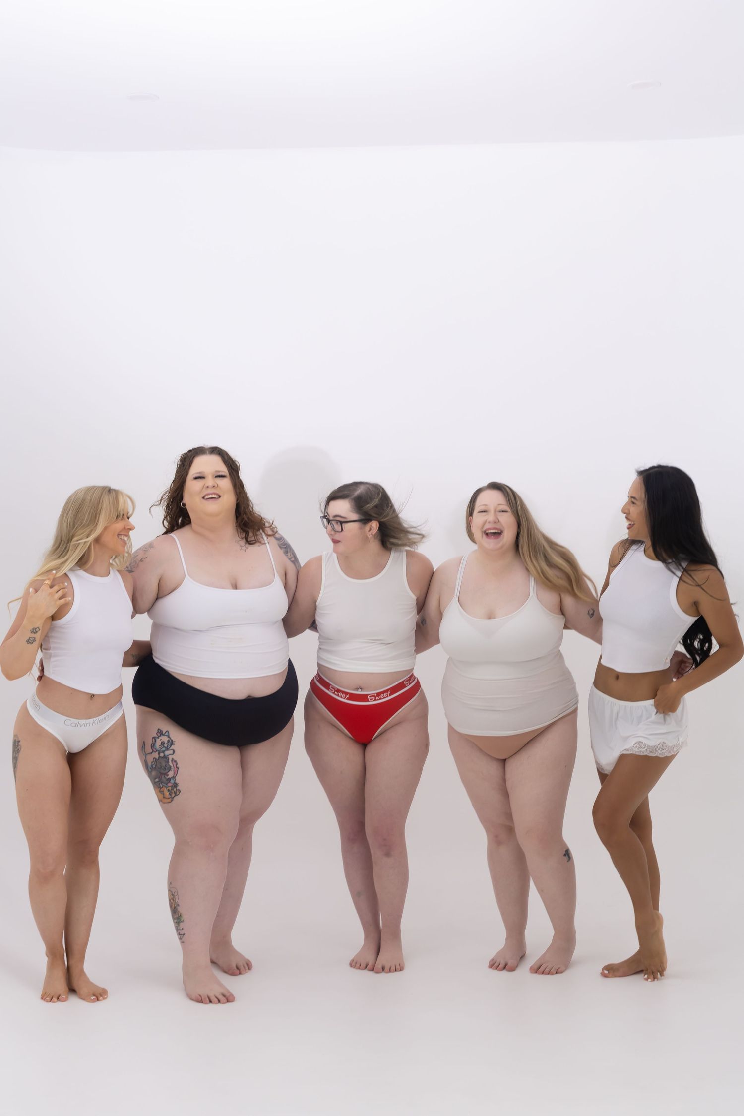 A diverse group of women in white tops and underwear pose together in a body-positive photoshoot against a white backdrop.
