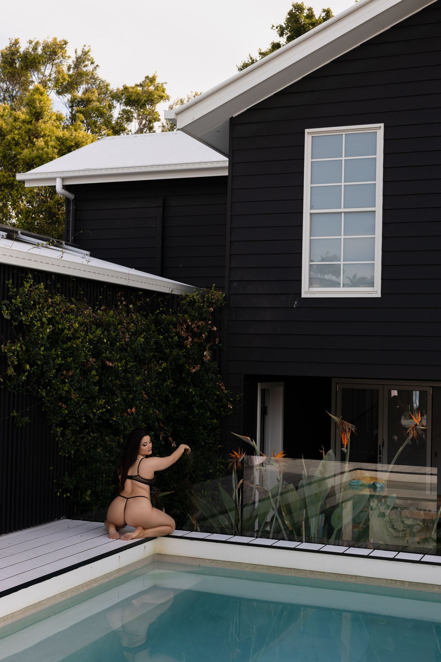 Modern black house with white trim and glass-paneled swimming pool surrounded by lush greenery in backyard setting.