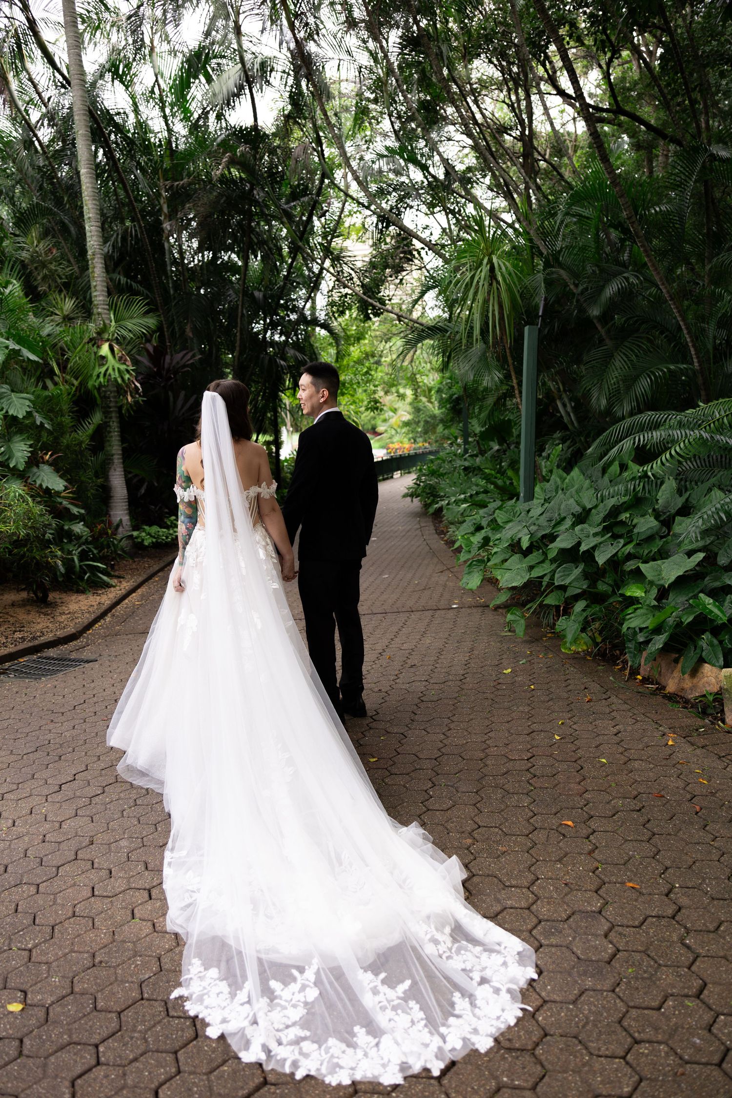 Bride and groom walking hand in hand on a brick path surrounded by lush tropical greenery, bride wearing a long lace veil.