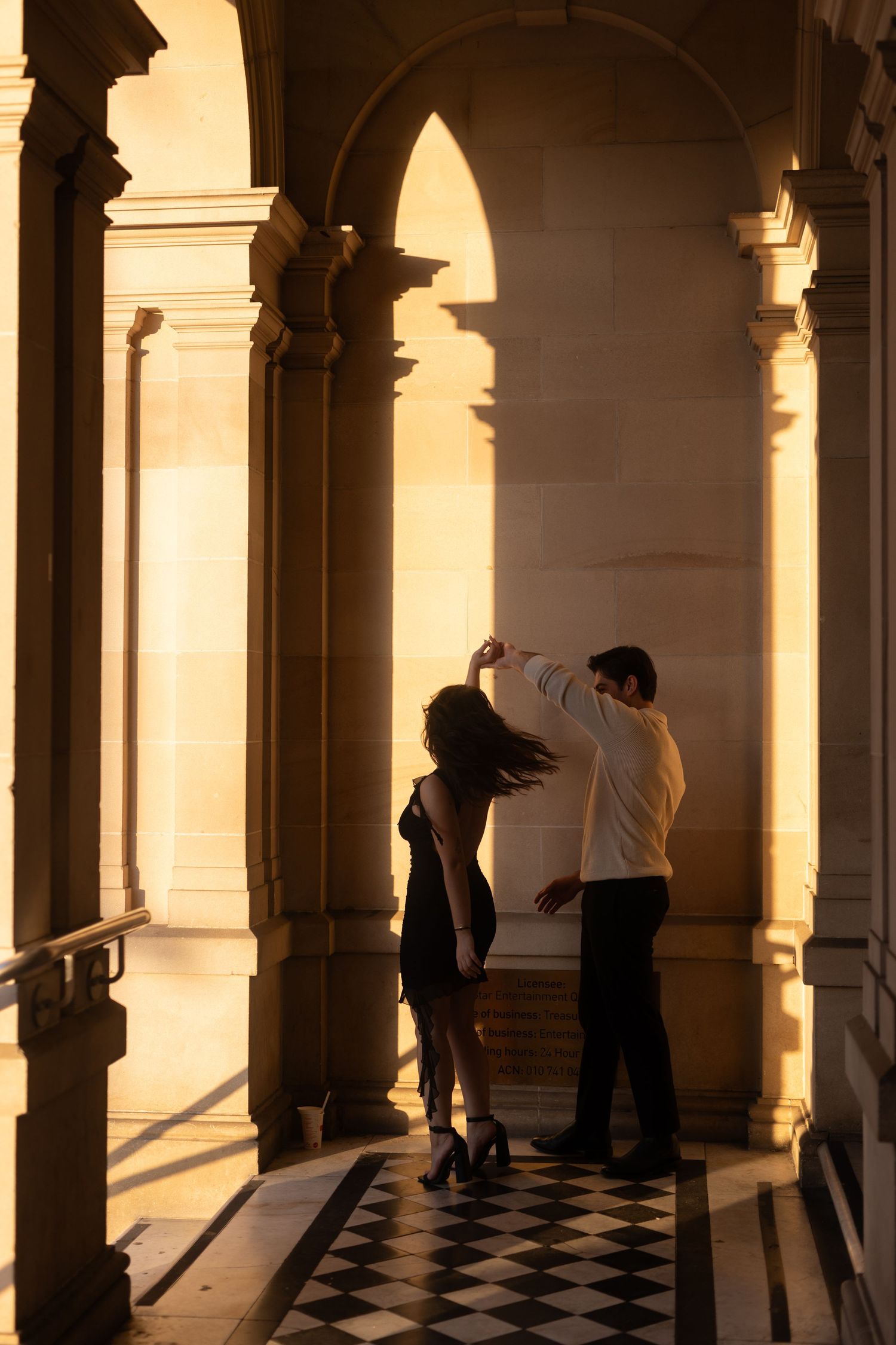 Silhouettes of people walking through a columned hallway with checkered floor in warm sunlight.