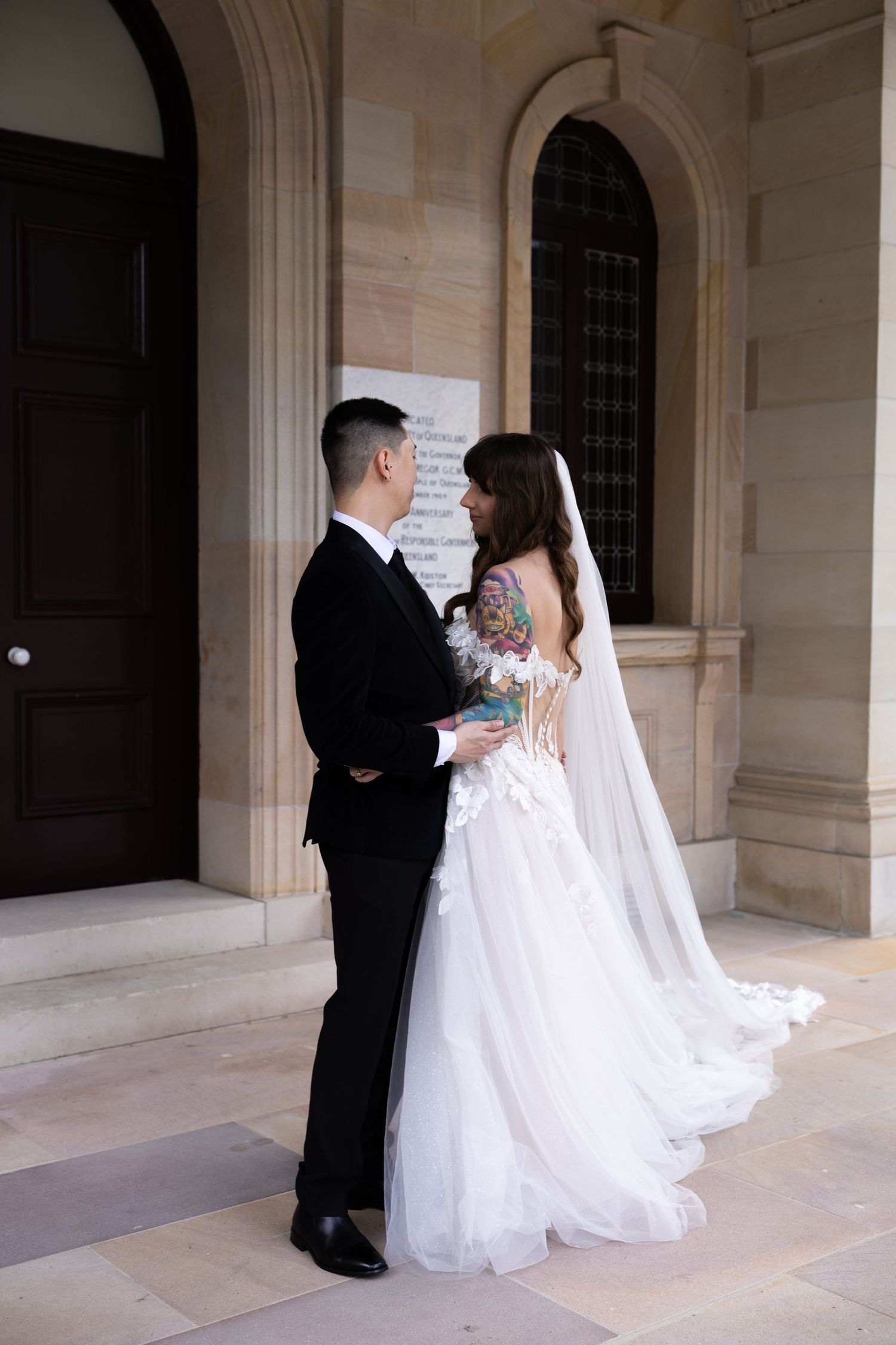 Bride and groom share a romantic moment outside a stone building, bride wearing a backless white gown with floral tattoos.