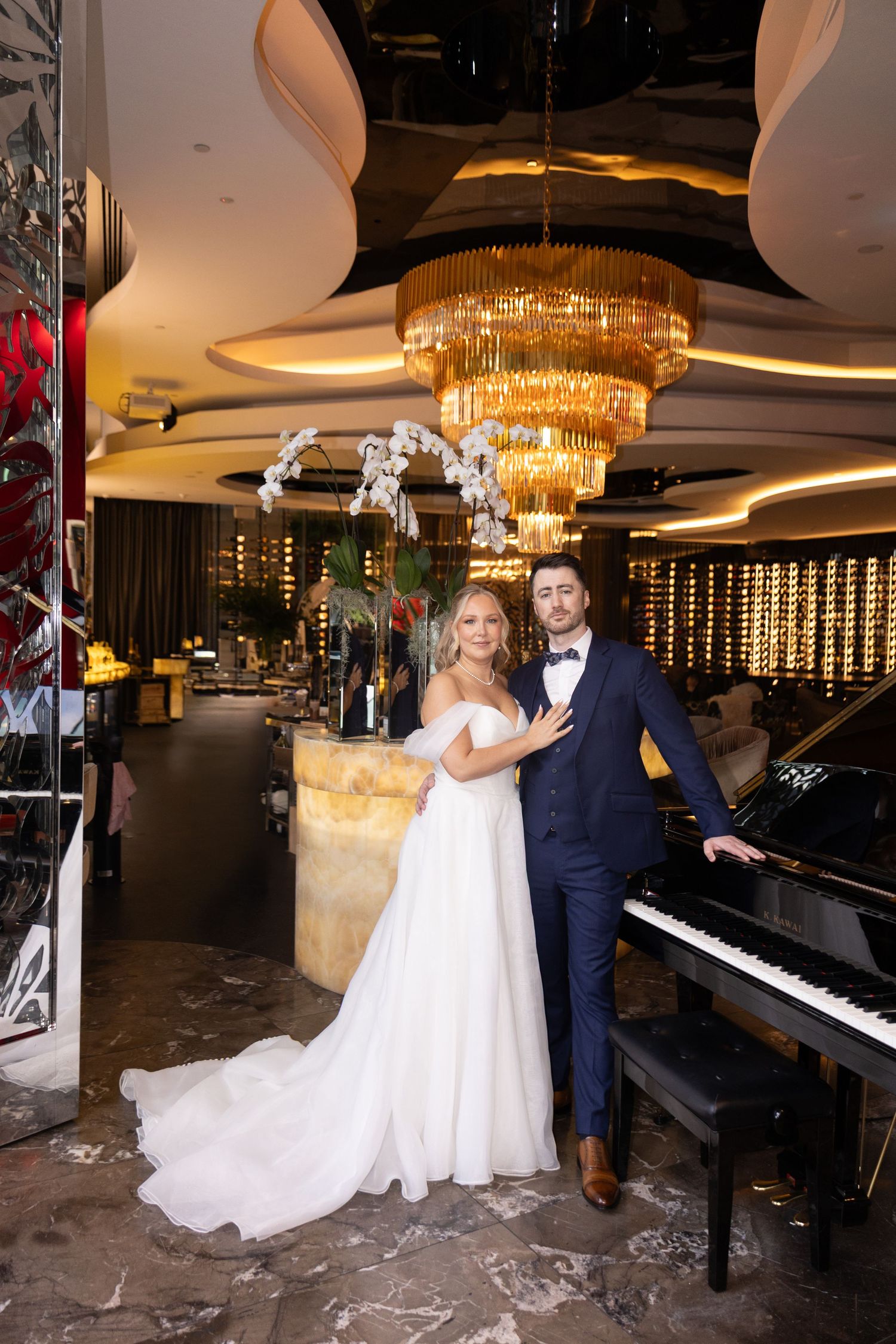 Bride in white gown and groom in navy suit pose by grand piano under gold chandelier in elegant venue.