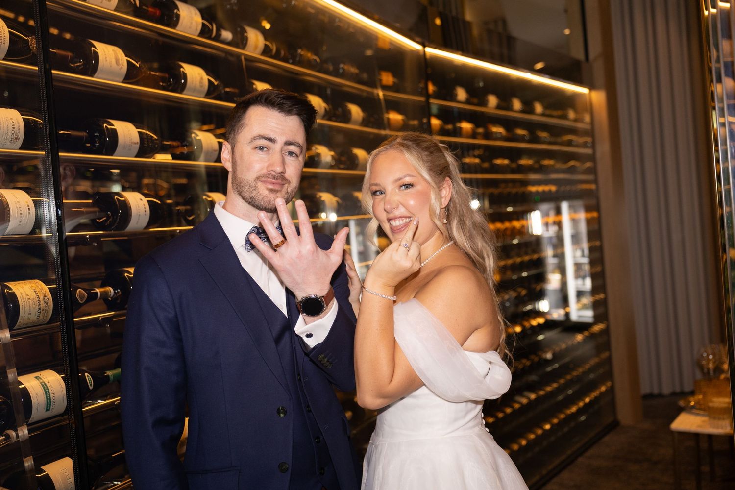 Bride and groom posing in a wine cellar, showing off wedding rings, with rows of wine bottles in the background.