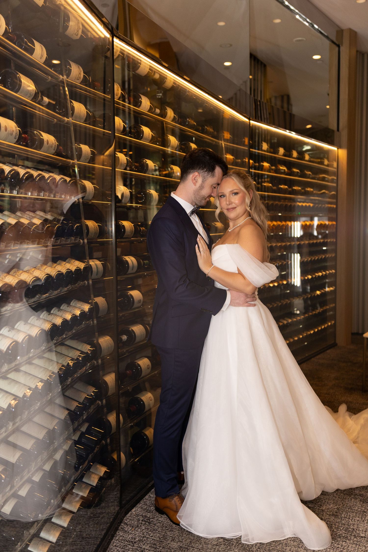 Bride and groom embracing in front of illuminated wine rack wall, bride in off-shoulder gown, groom in navy tuxedo.