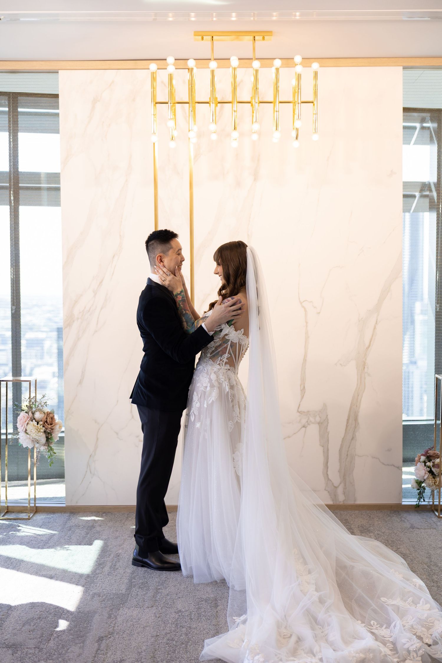 Bride and groom share a tender moment before a marble backdrop under a gold chandelier in an elegant wedding venue.