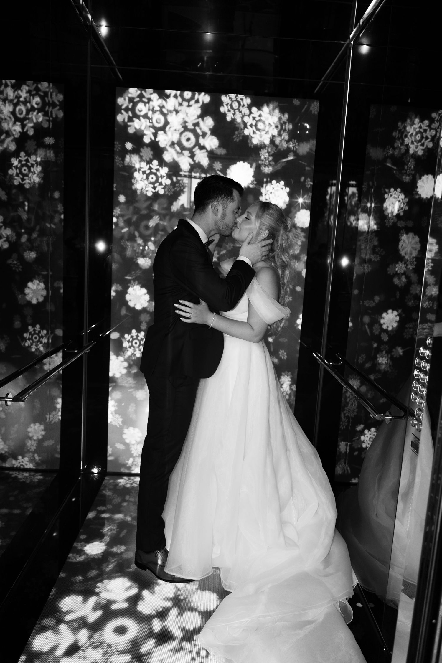 Black and white photo of a bride and groom sharing a kiss, surrounded by floral light projections.