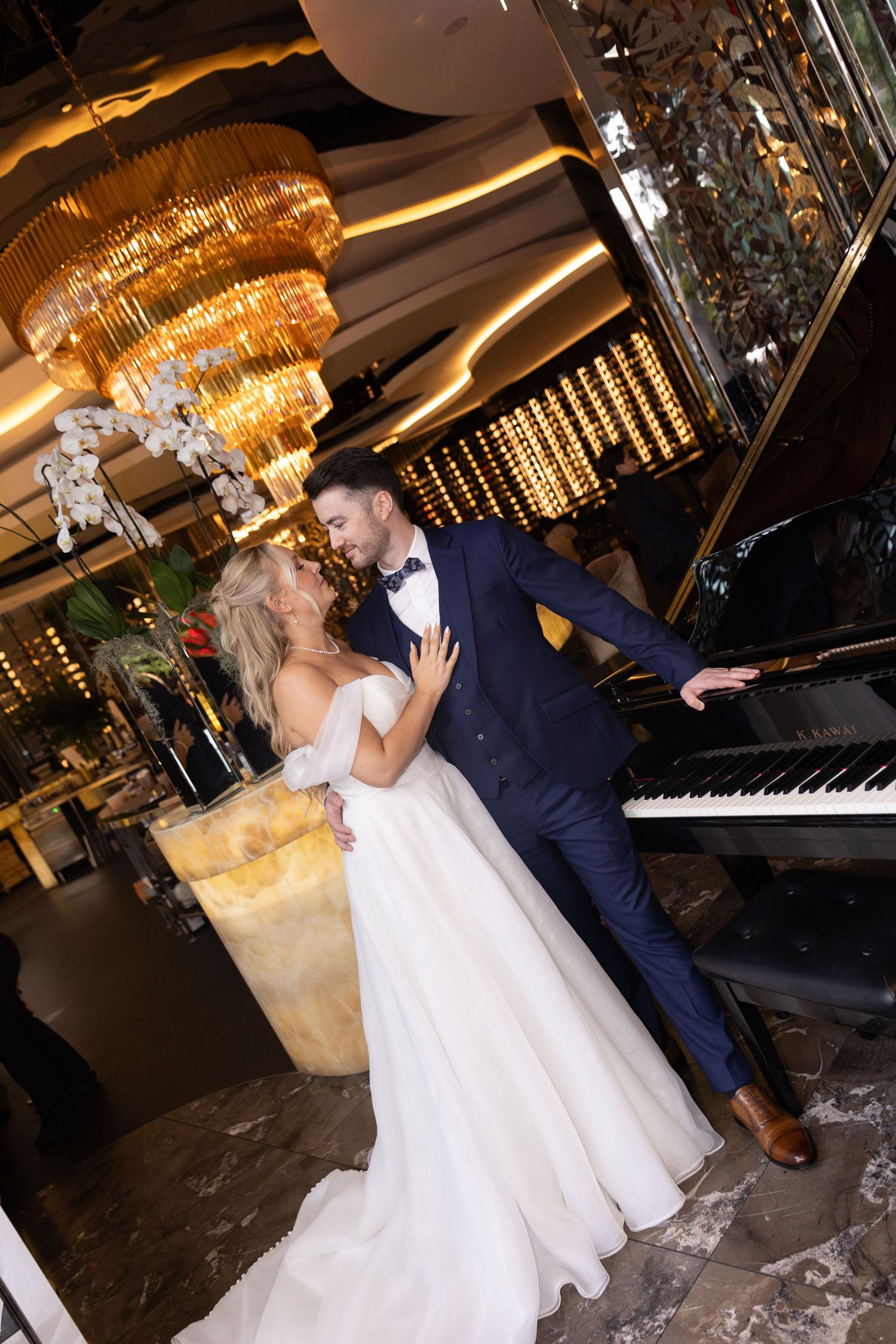Bride and groom share a romantic moment by a grand piano in an elegant venue with a gold chandelier.