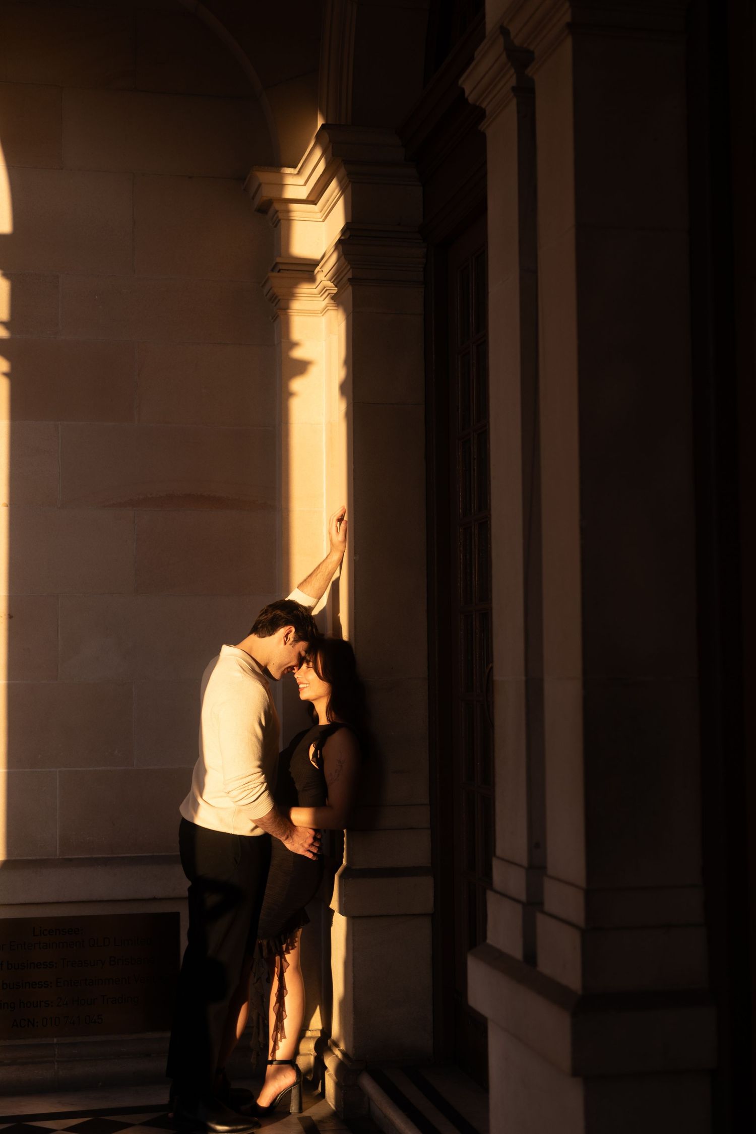 Silhouetted couple embraces in a dramatic colonnade hallway with sunlit pillars creating a moody architectural atmosphere.