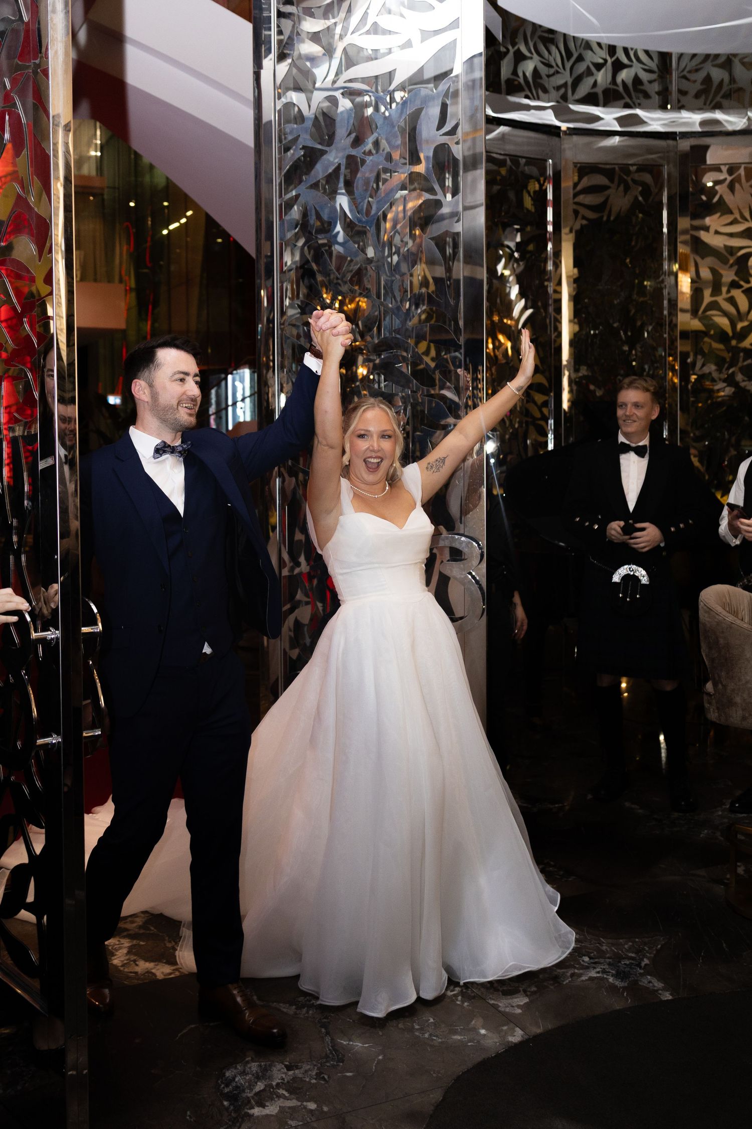 Joyful newlyweds raise hands in celebration as bride in white gown and groom in navy suit exit wedding venue.