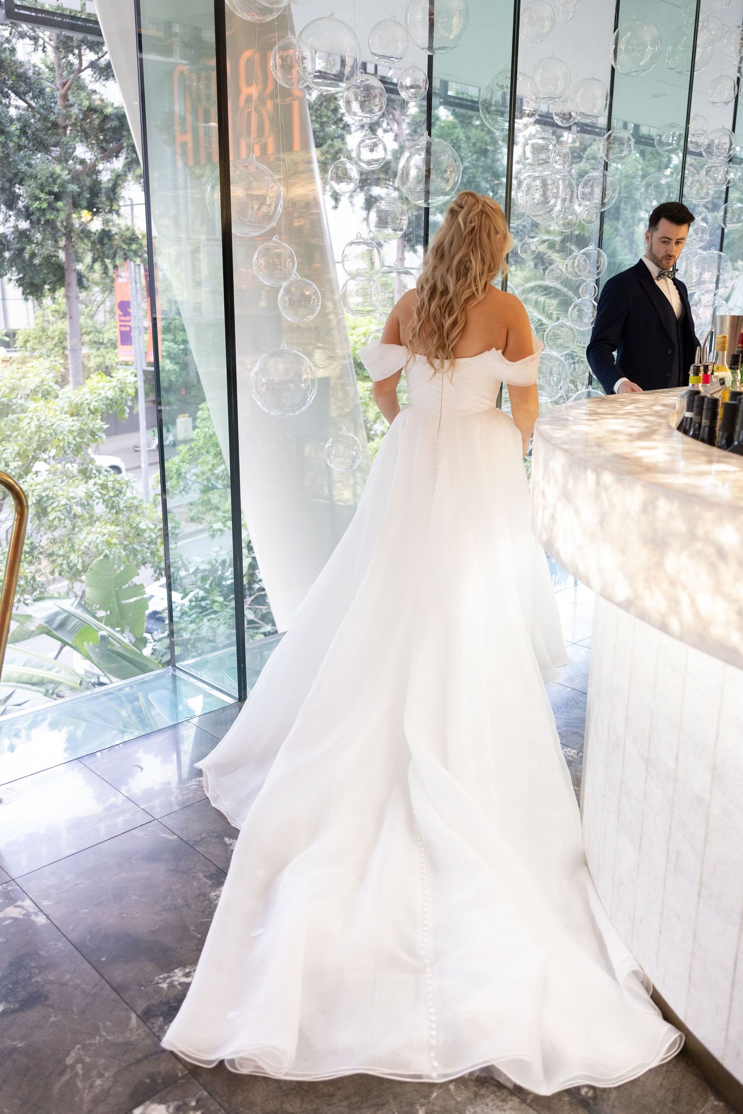 Bride in elegant off-shoulder white ball gown with long train stands near glass wall, groom in navy suit at bar.
