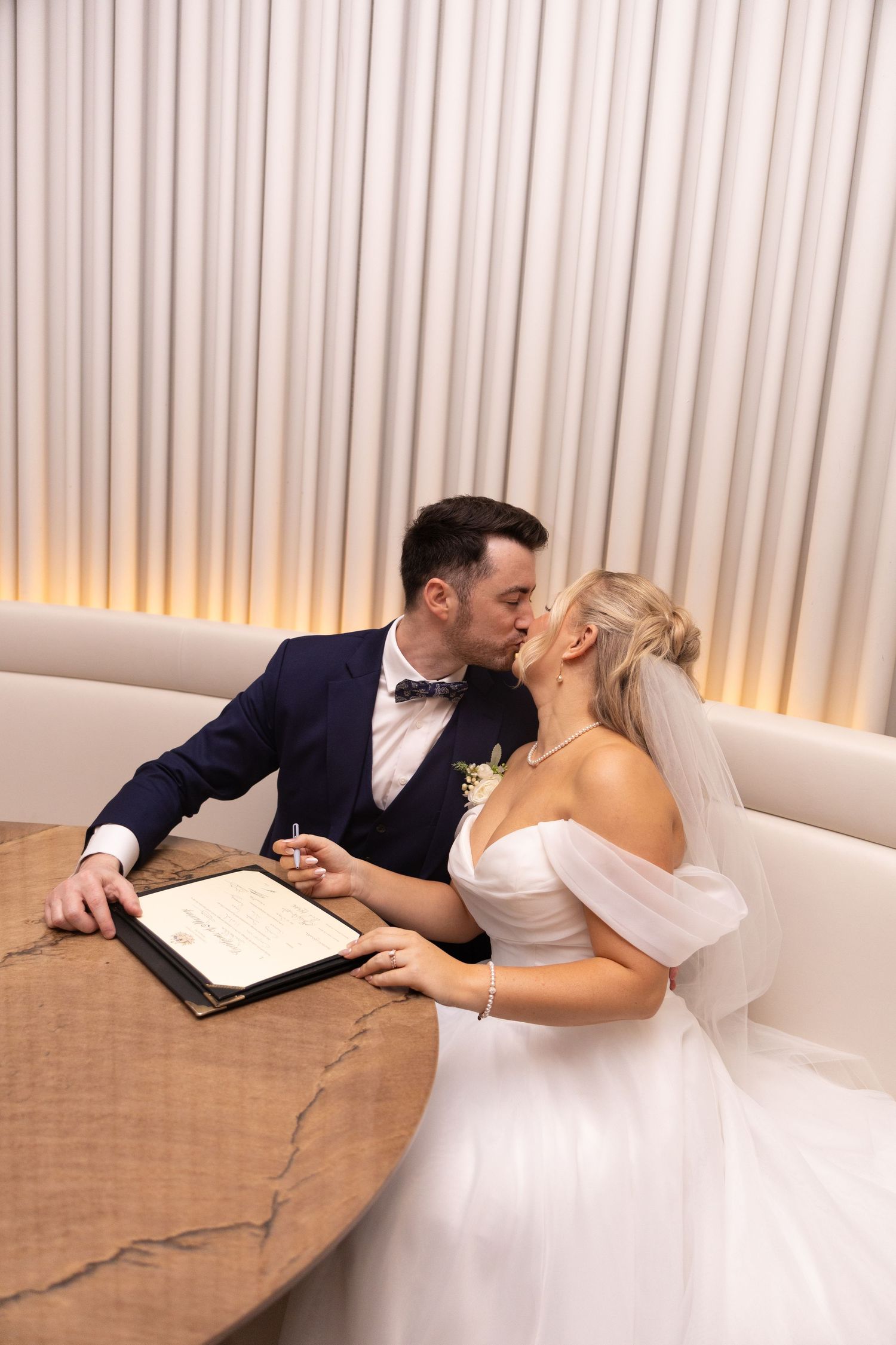 Bride and groom share a kiss while signing their marriage certificate at a wooden table during their wedding ceremony.