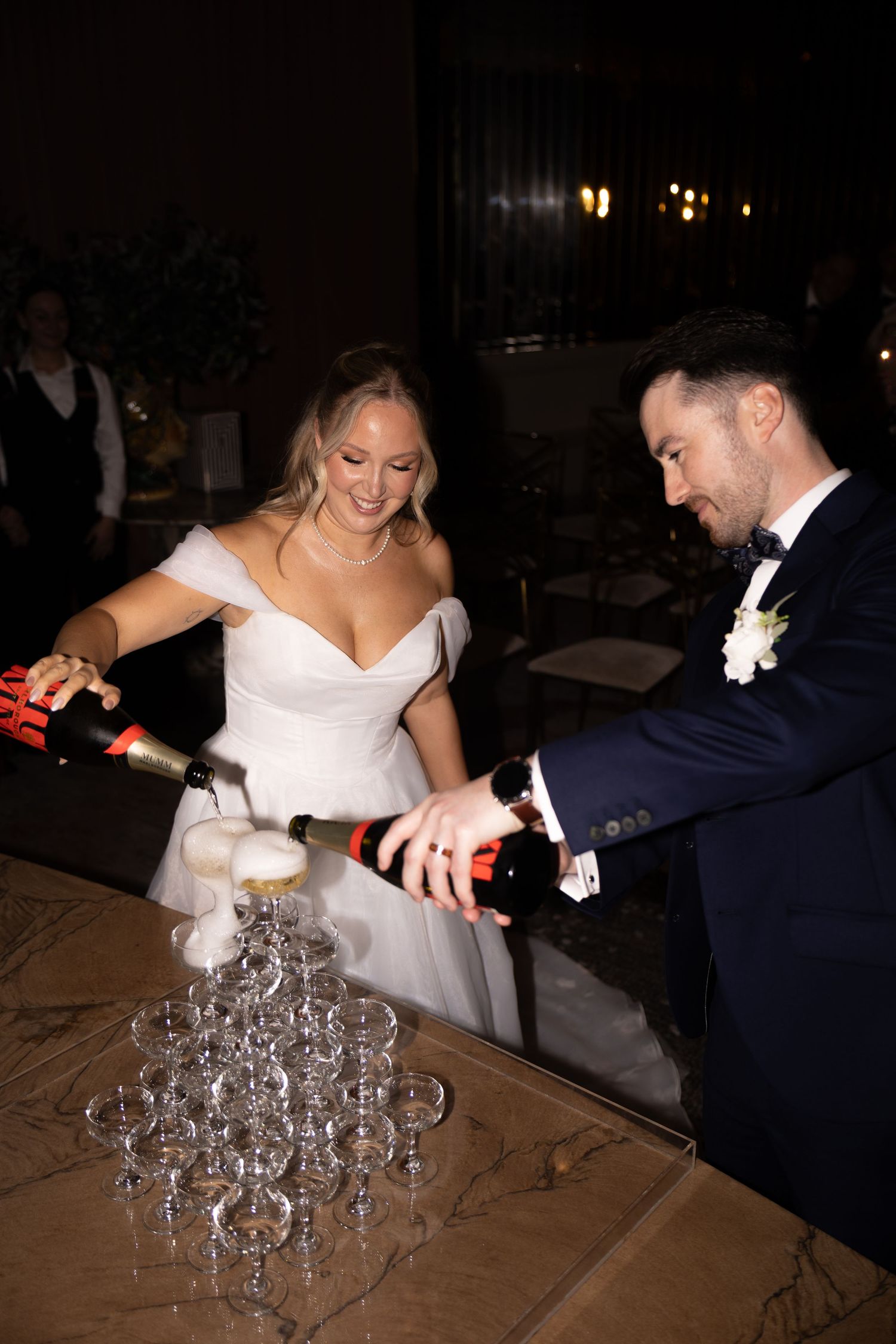Bride and groom pouring champagne tower at wedding reception in elegant dark venue.