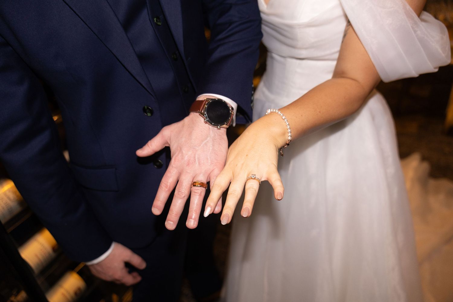 Bride and groom displaying wedding rings on outstretched hands, groom wearing navy suit and watch.