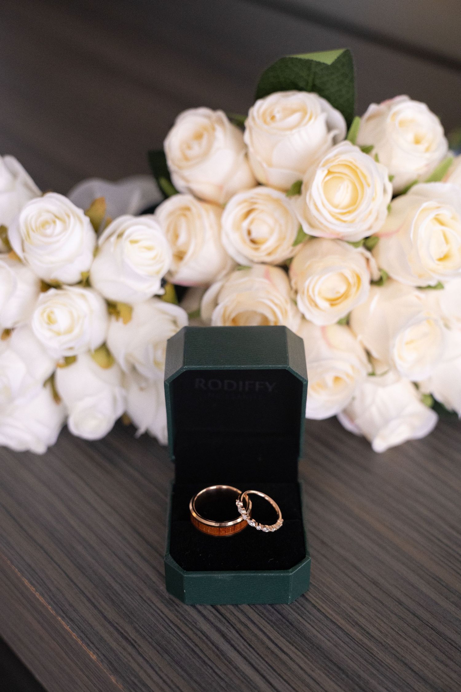 Two gold wedding rings in a green Rodifty box beside a bouquet of white roses on a wooden surface.