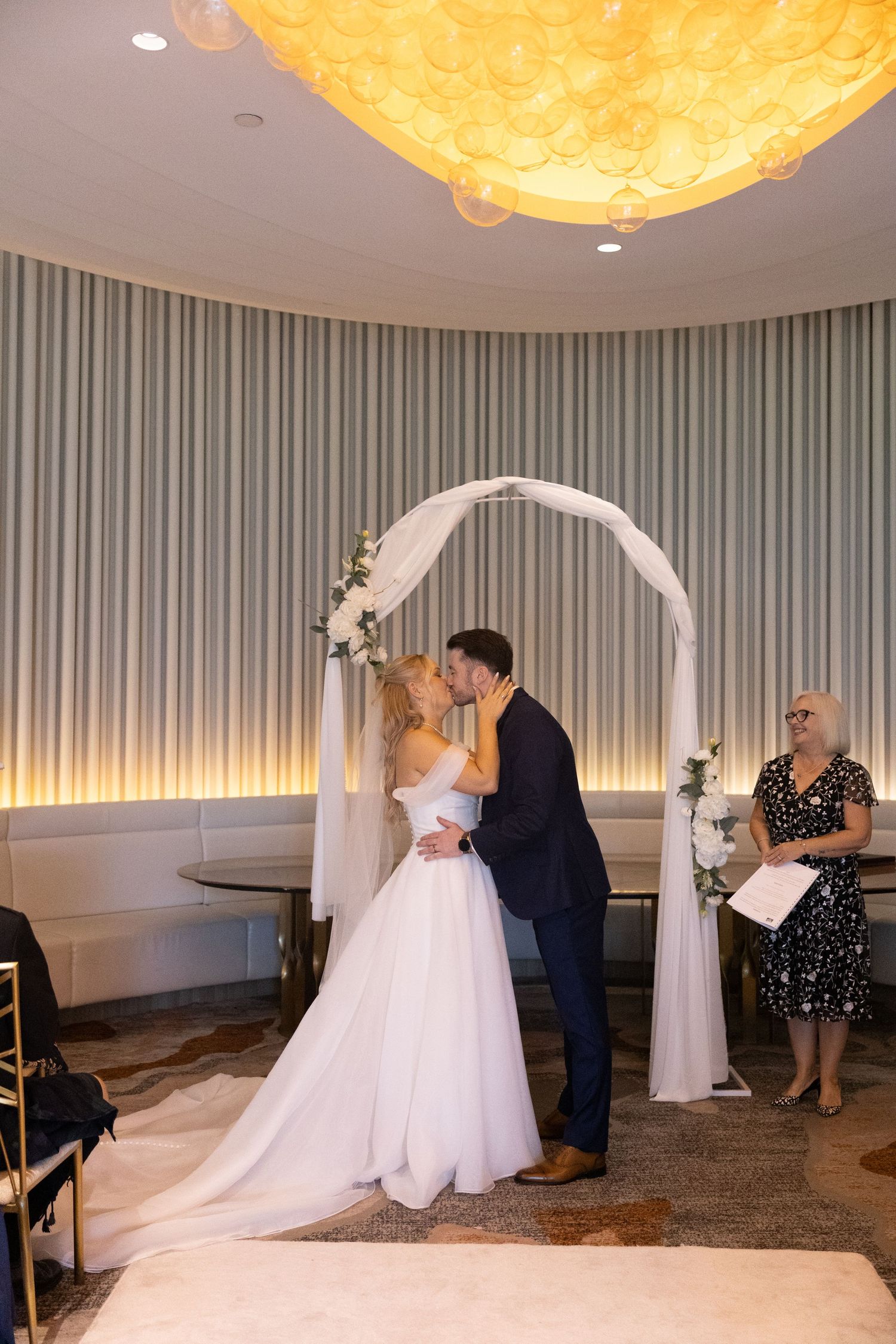 Bride and groom share first kiss under white floral arch at elegant indoor wedding ceremony.
