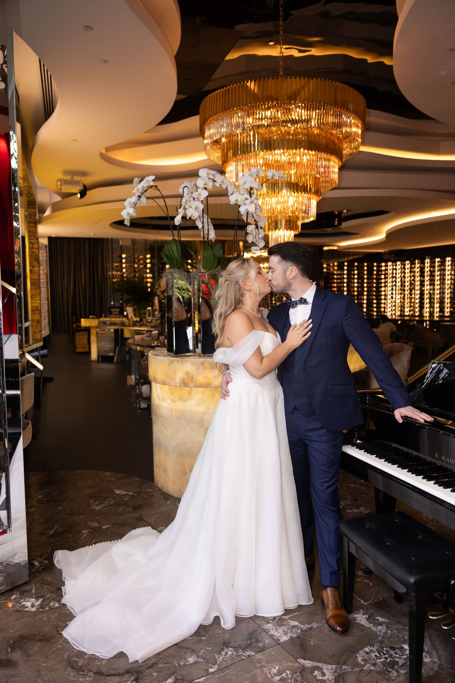 A bride and groom share a romantic kiss in an elegant venue with a grand chandelier and piano.