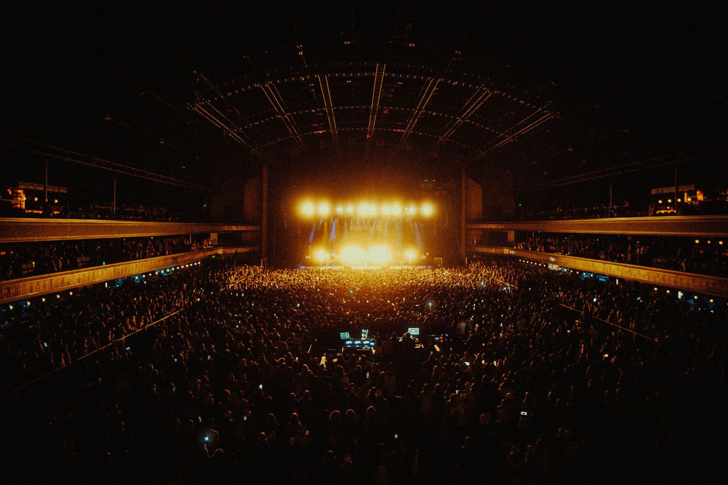 Packed concert arena with massive glowing stage lights illuminating thousands of fans in a dark indoor venue.