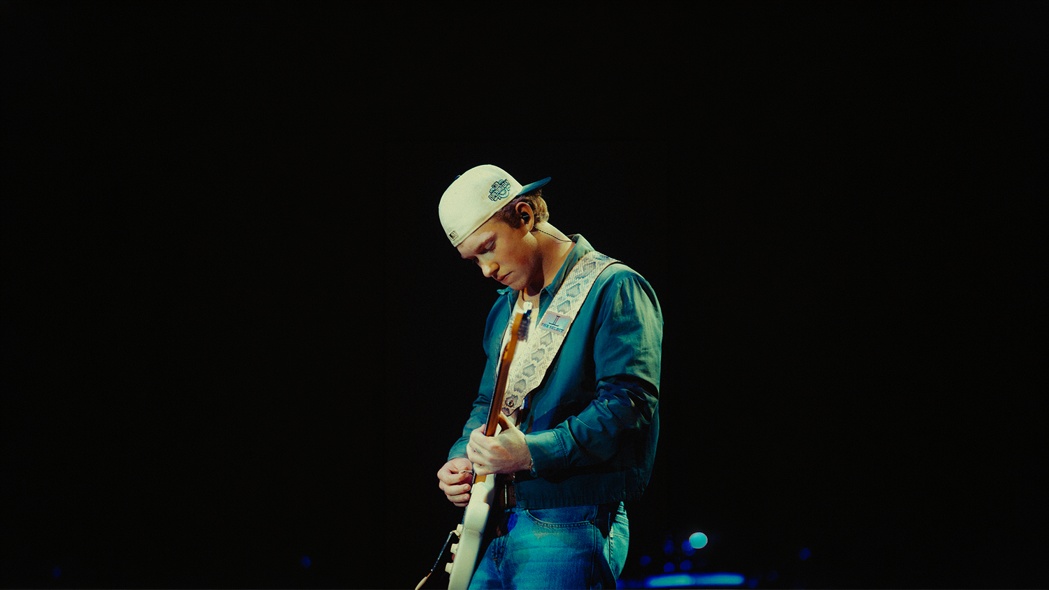 Ty Myers playing electric guitar on stage under spotlight, wearing white cap and blue jacket in dark concert setting.
