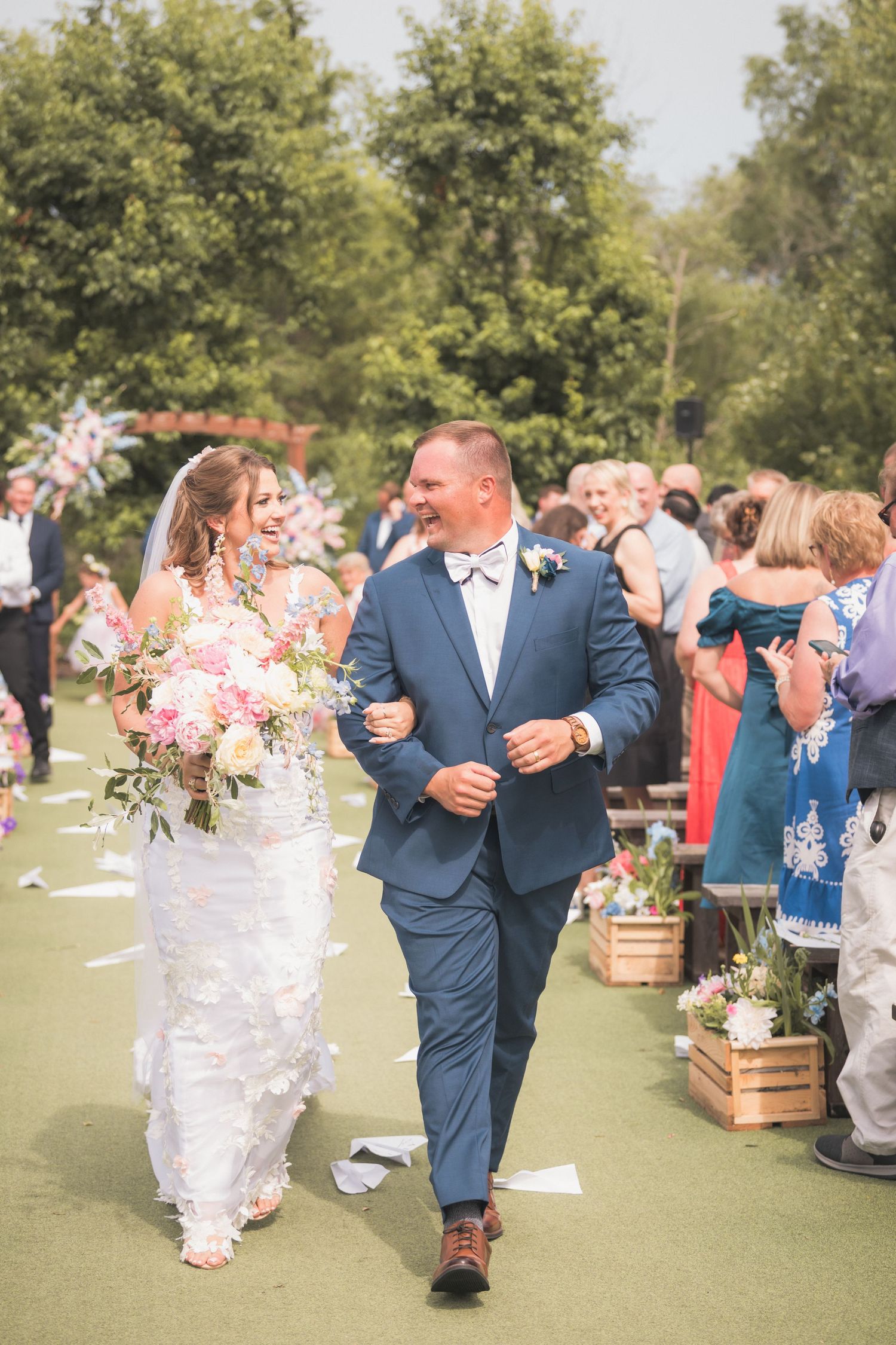 Bride and groom walk down the aisle smiling, bride holding pink bouquet, groom in blue suit, outdoor wedding ceremony.