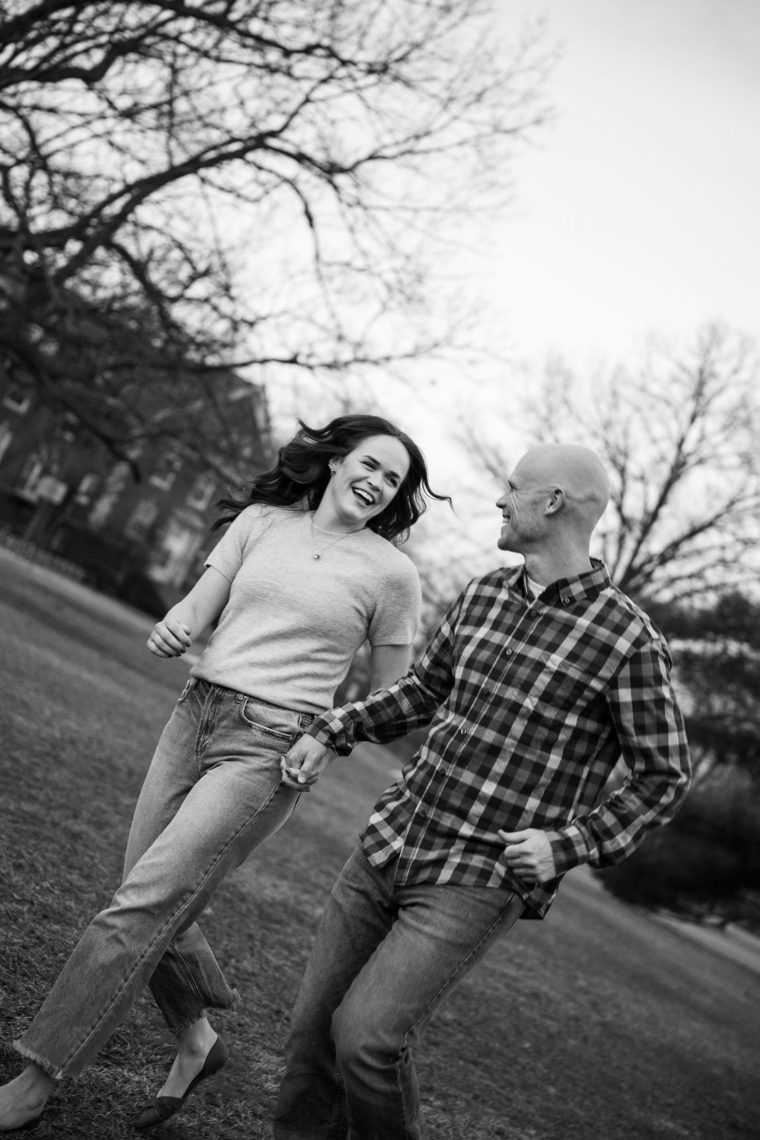 Black and white photo of a laughing couple holding hands and walking outdoors near bare trees.