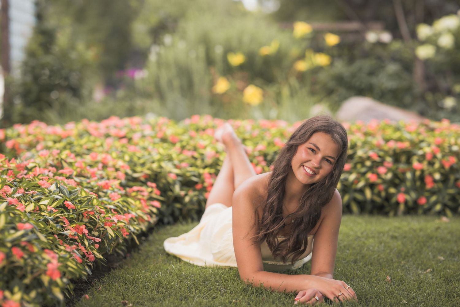 Young woman in white dress lying on green grass smiling, surrounded by colorful blooming flowers in a garden.