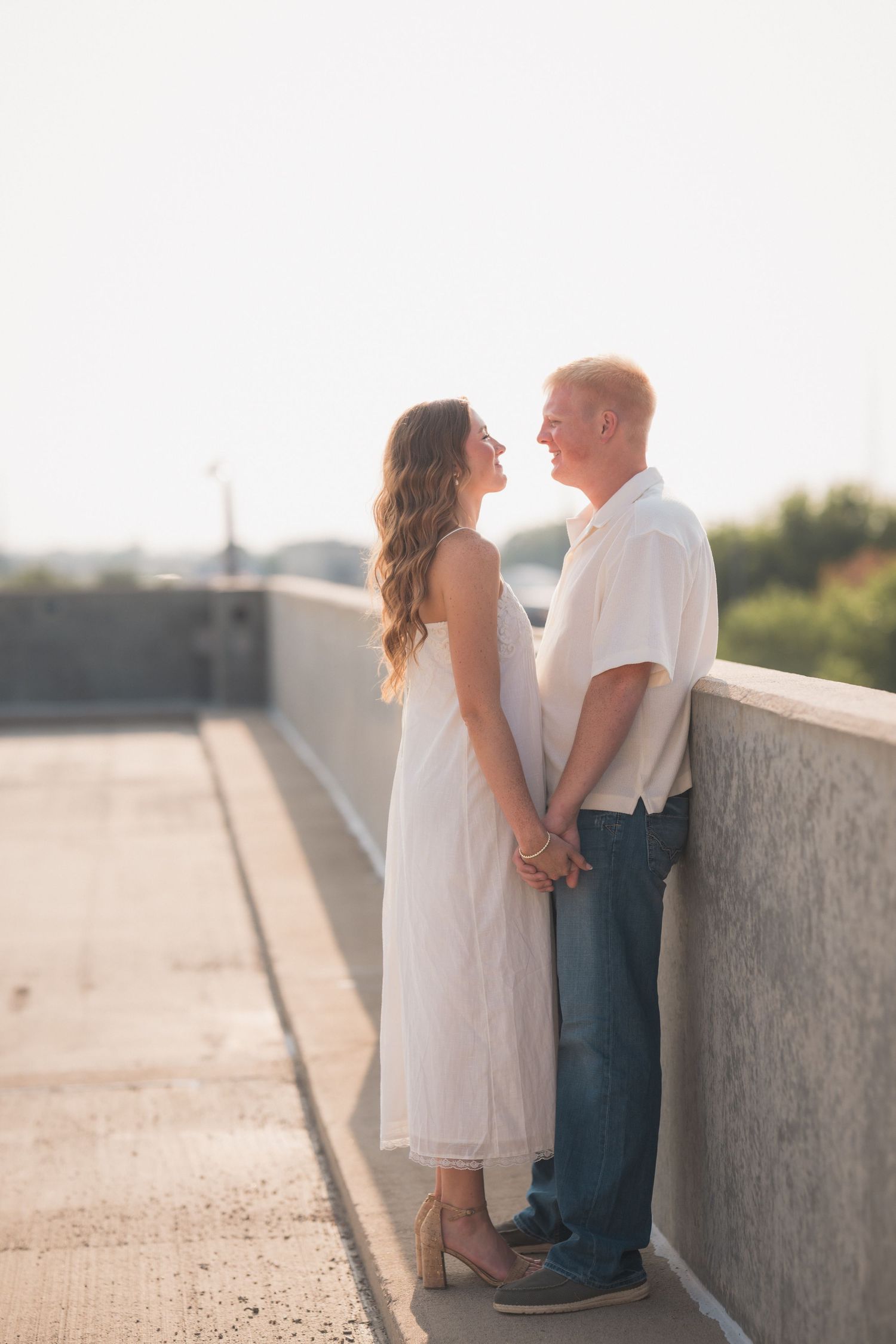 Couple holding hands and gazing at each other on a bridge, woman in white dress, man in white shirt and jeans.