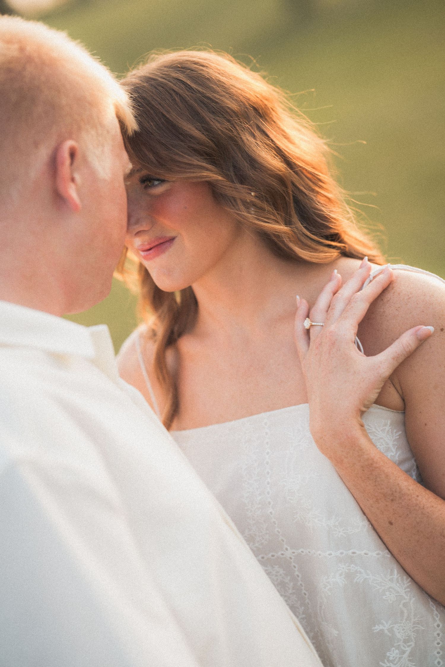 Bride in white dress gazes at groom with engagement ring visible on her shoulder during golden hour outdoor wedding photo.