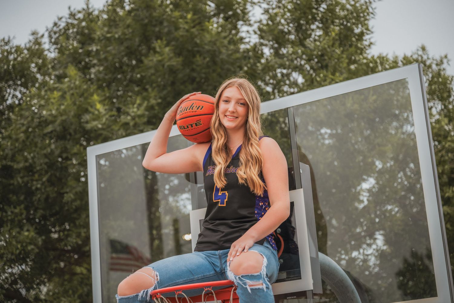 Blonde girl in basketball jersey sitting on hoop backboard holding basketball outdoors.