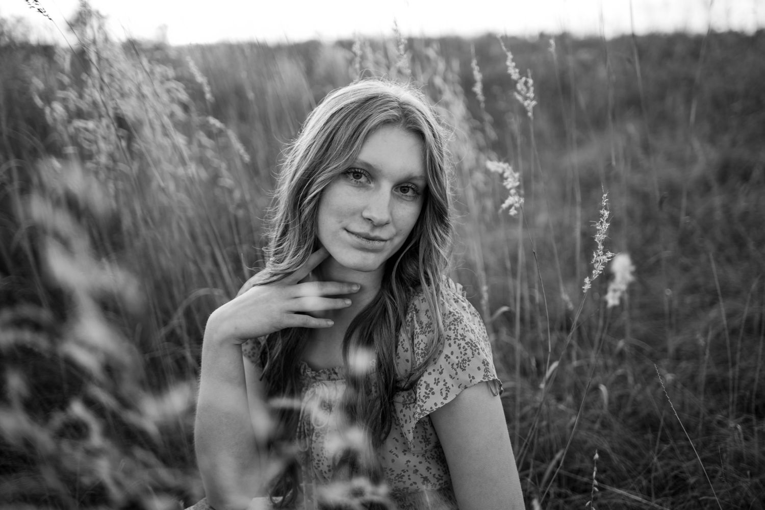 Close-up black and white portrait of young woman sitting in tall grass field at golden hour.
