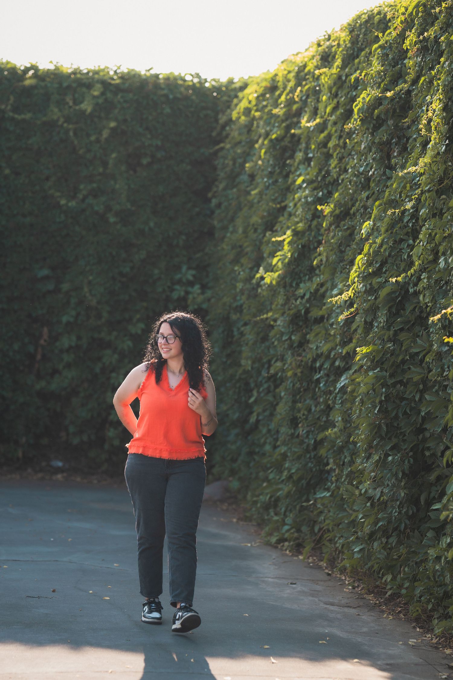 Woman in orange top and gray pants walking between tall green hedges on a sunny day.