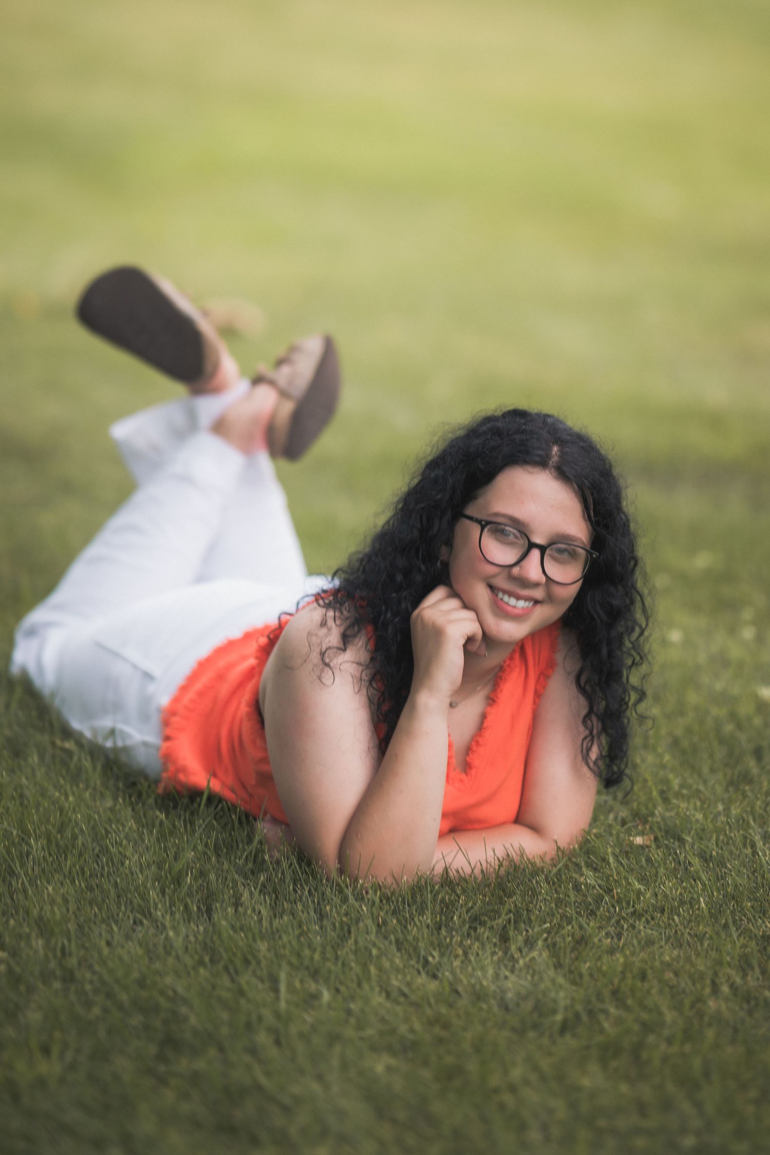 A smiling woman with curly hair and glasses lying on green grass wearing an orange top and white pants.
