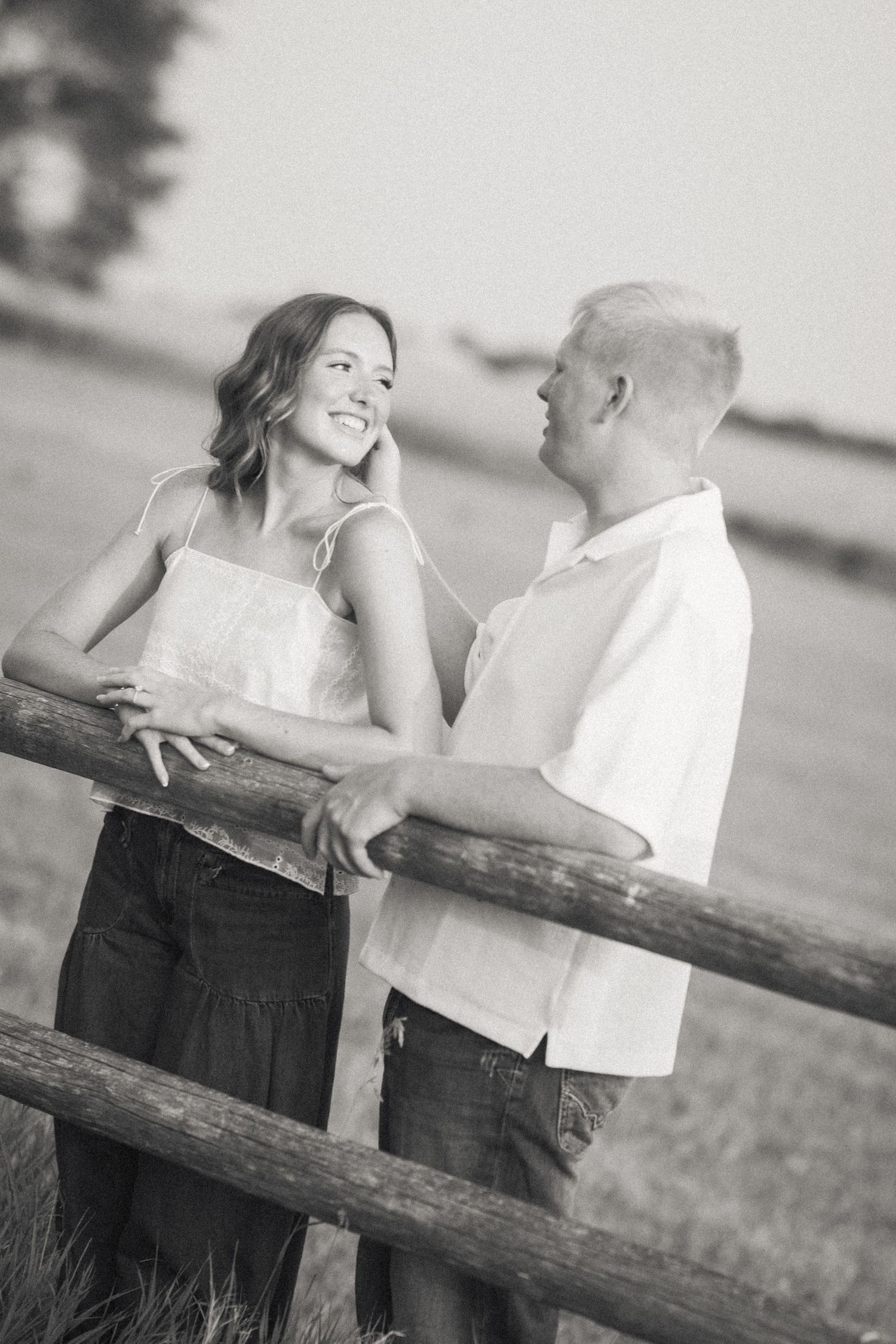 Black and white photo of a smiling couple leaning on a wooden fence in a rural field setting.