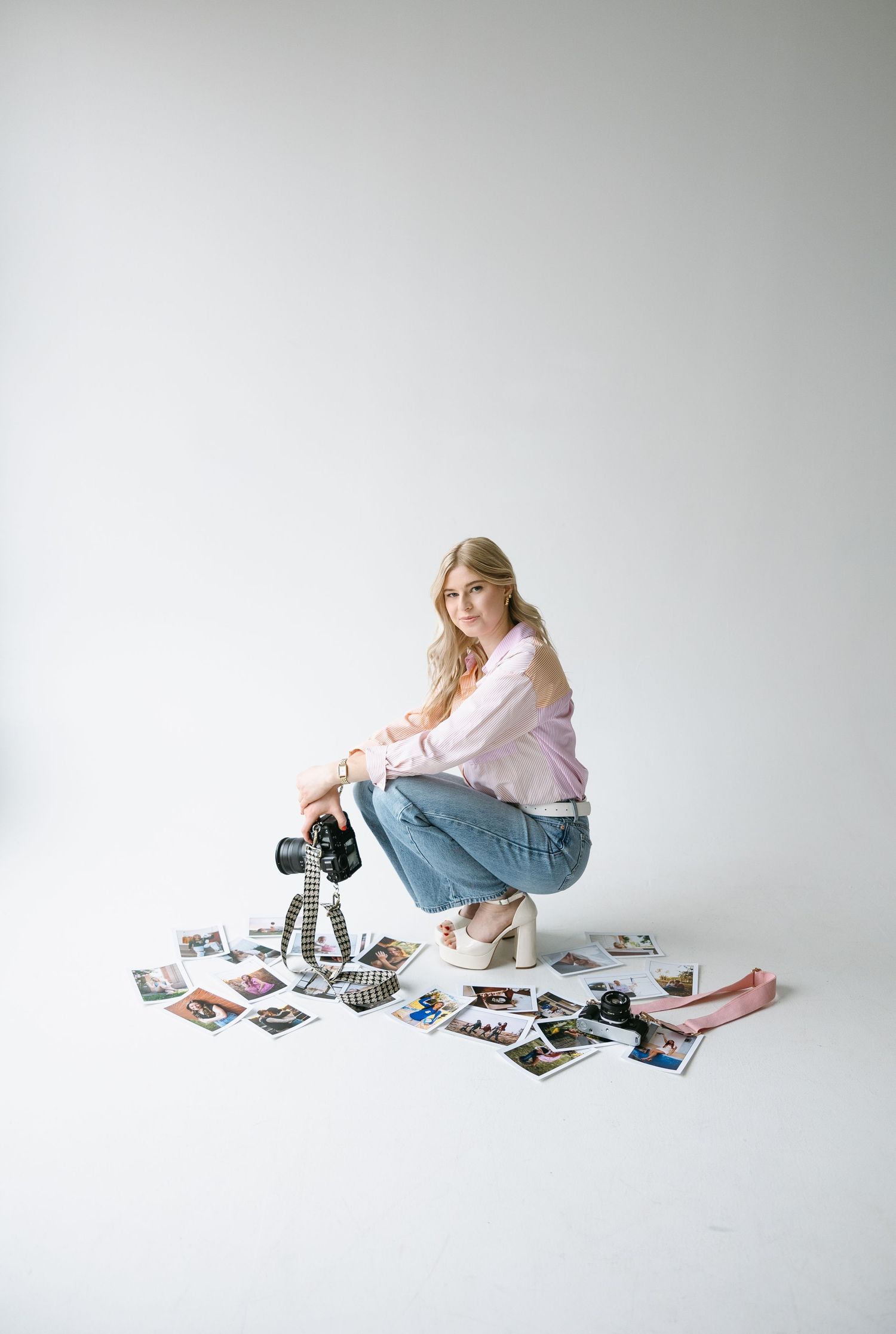Female photographer crouching with DSLR camera surrounded by printed photos on white studio floor.