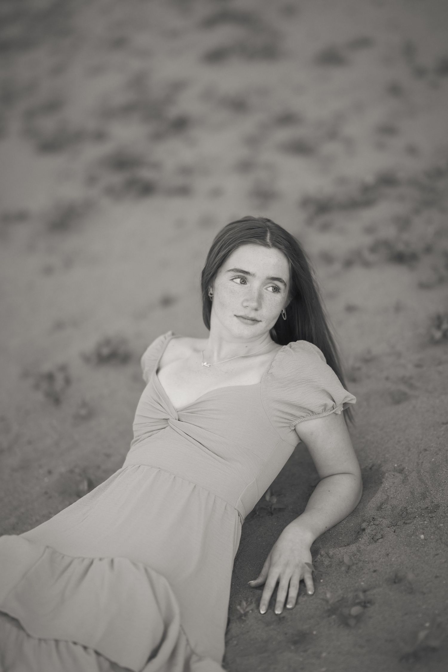 Black and white photo of a young woman in a dress sitting on sandy beach, looking up with a serene expression.