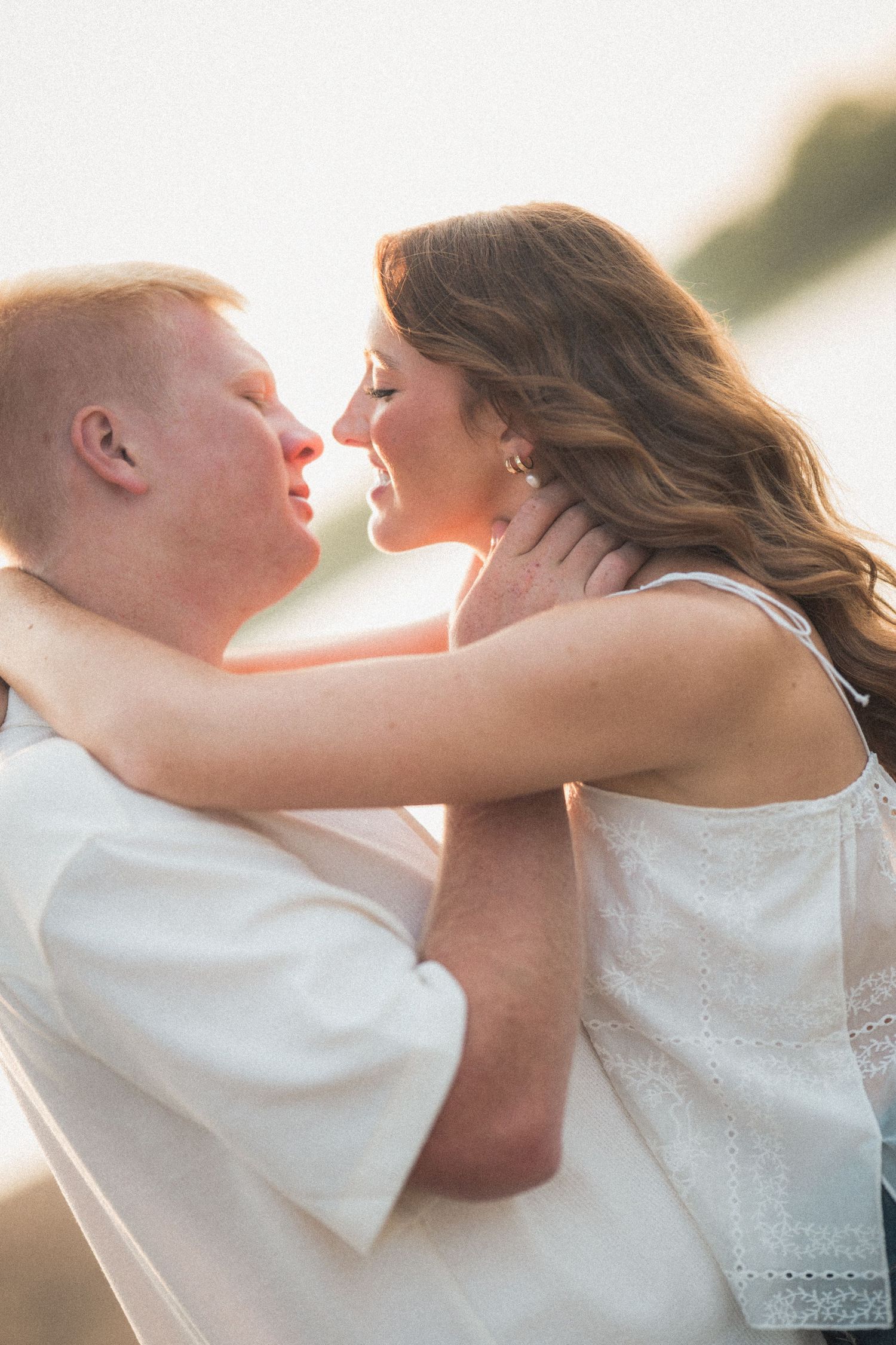 A couple in white clothing shares a romantic moment at golden hour, nearly kissing outdoors near water.