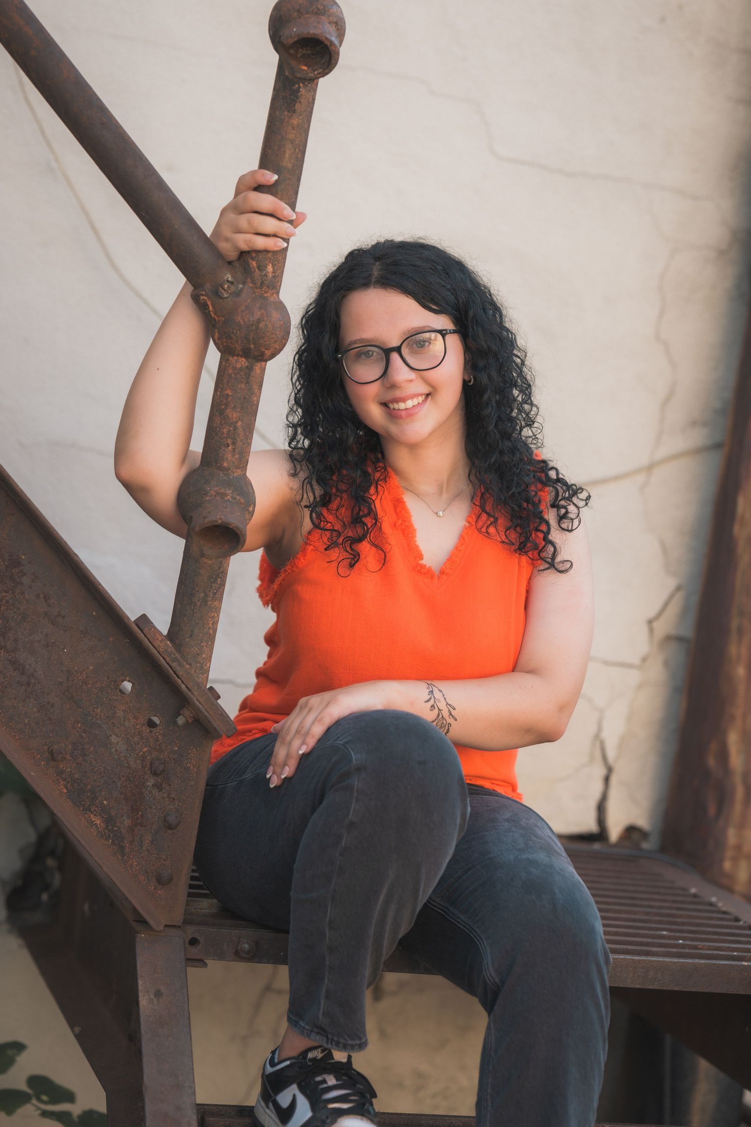 A smiling woman with curly dark hair and glasses sits on rustic metal stairs wearing an orange sleeveless top and gray jeans.