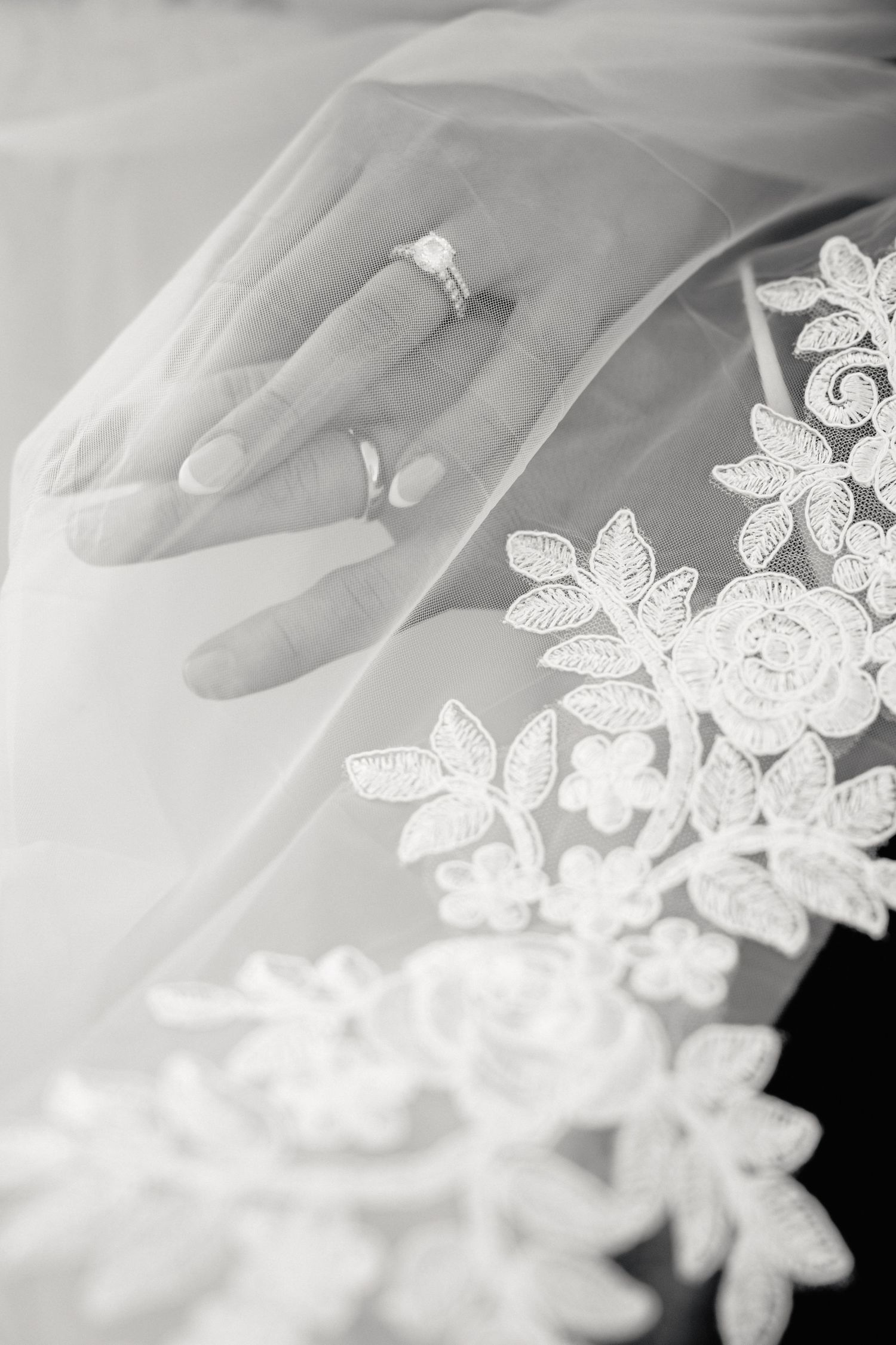 Black and white close-up of a bride's hands holding a lace veil, wearing an engagement ring.